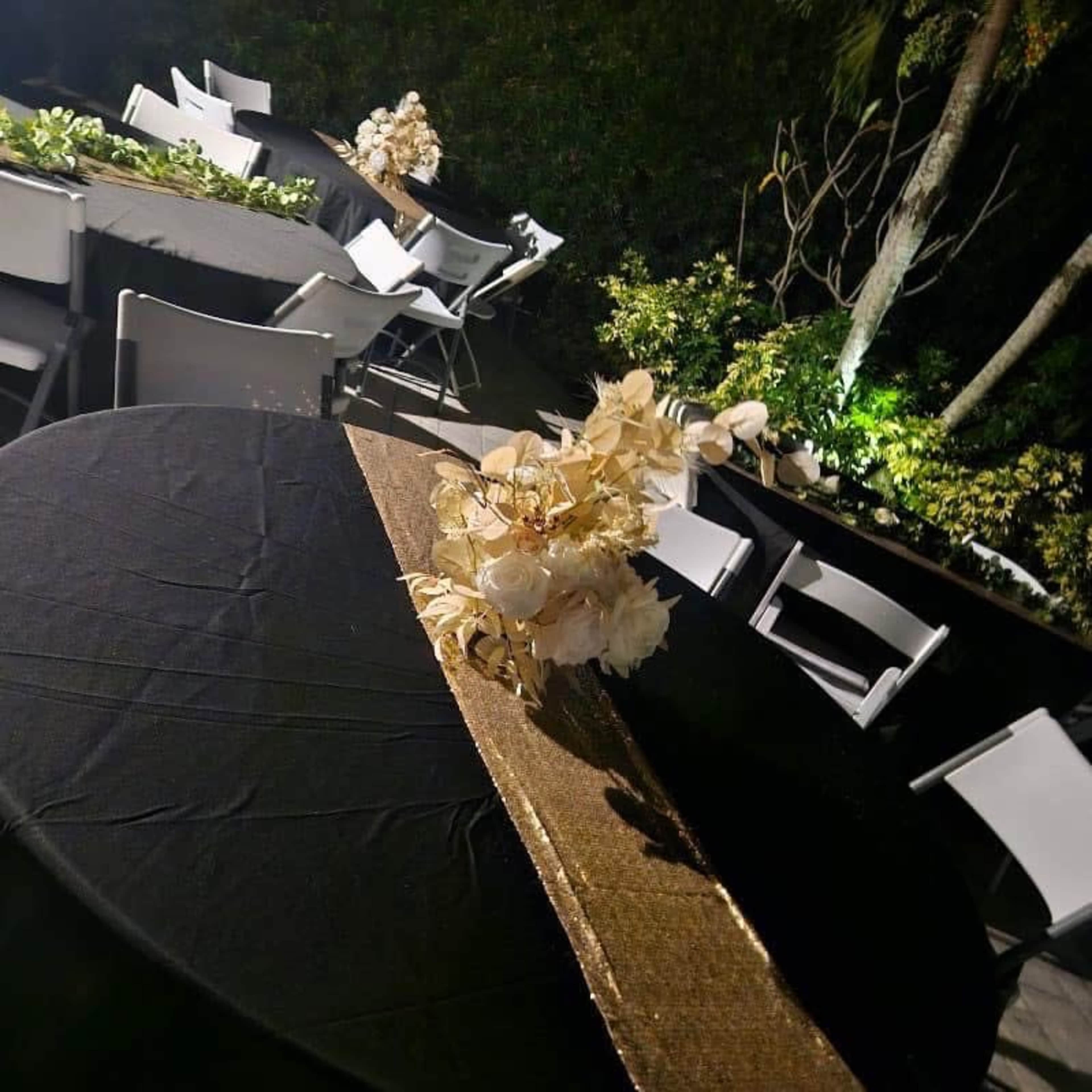 A table is set outdoors at night, decorated with a gold runner and floral arrangements, surrounded by empty chairs and illuminated by soft lighting.