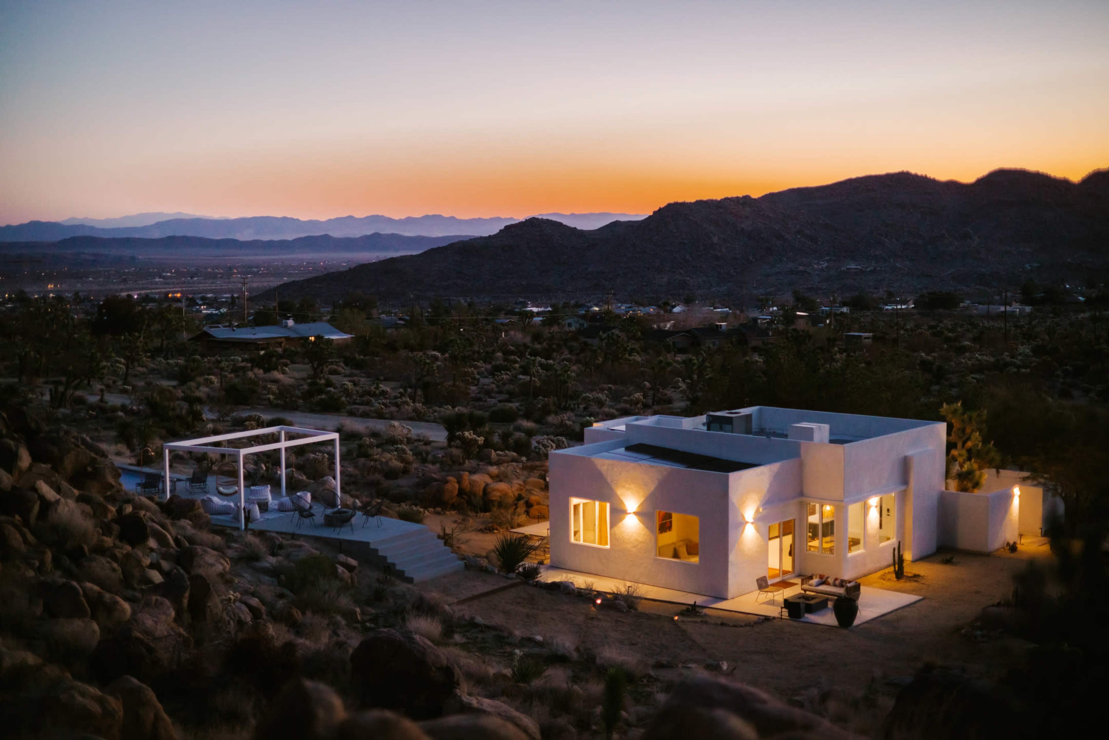 A modern white house is situated in a desert landscape at dusk, surrounded by mountains and rocky terrain.