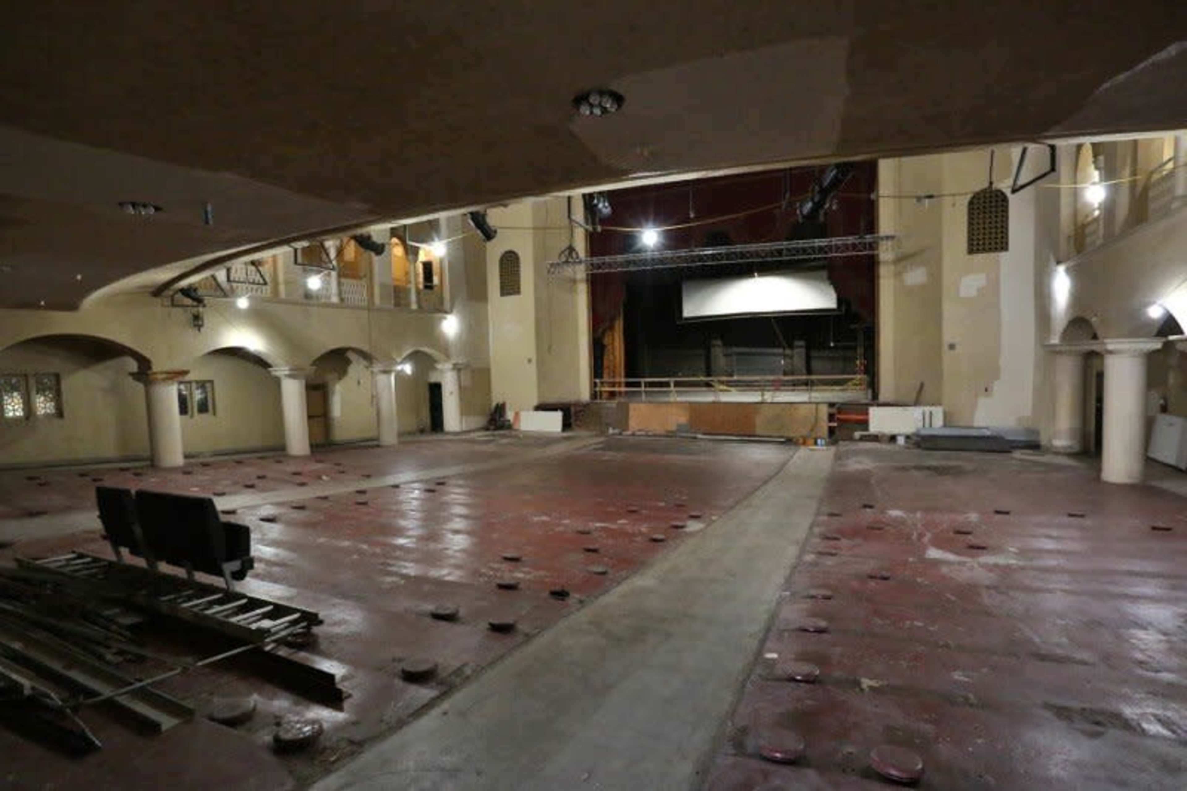 A partially renovated theater with exposed flooring, scattered construction materials, and a large stage at the front.