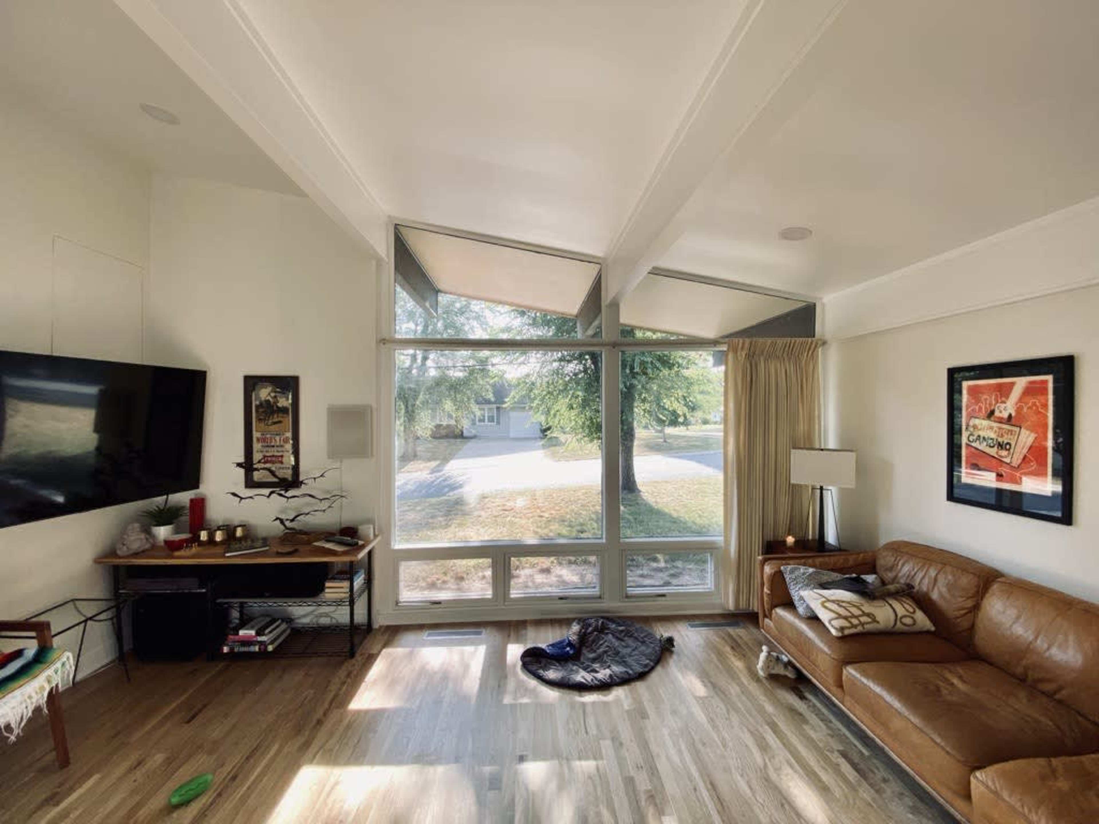 A spacious living room features a large window with geometric shades, a leather sofa, and a television mounted on the wall.