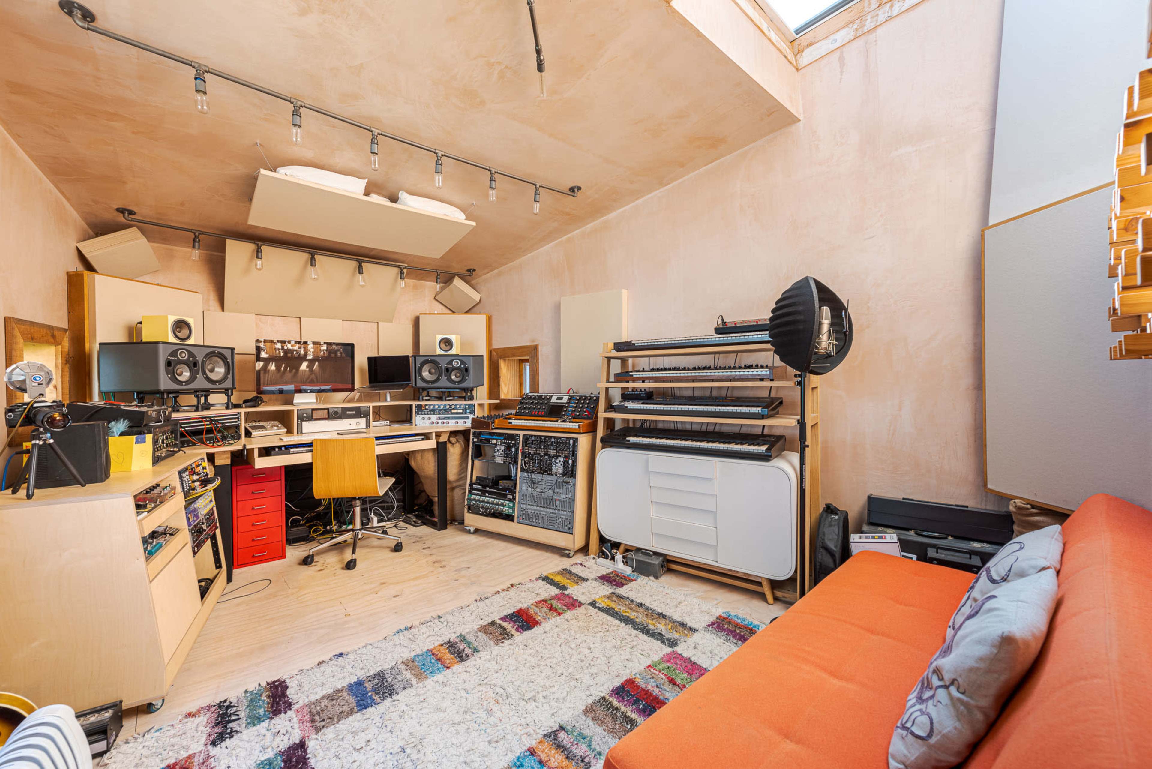 The image shows a well-equipped music studio featuring a central work station with audio equipment, a comfortable sofa, and textured walls with natural light coming from a skylight.