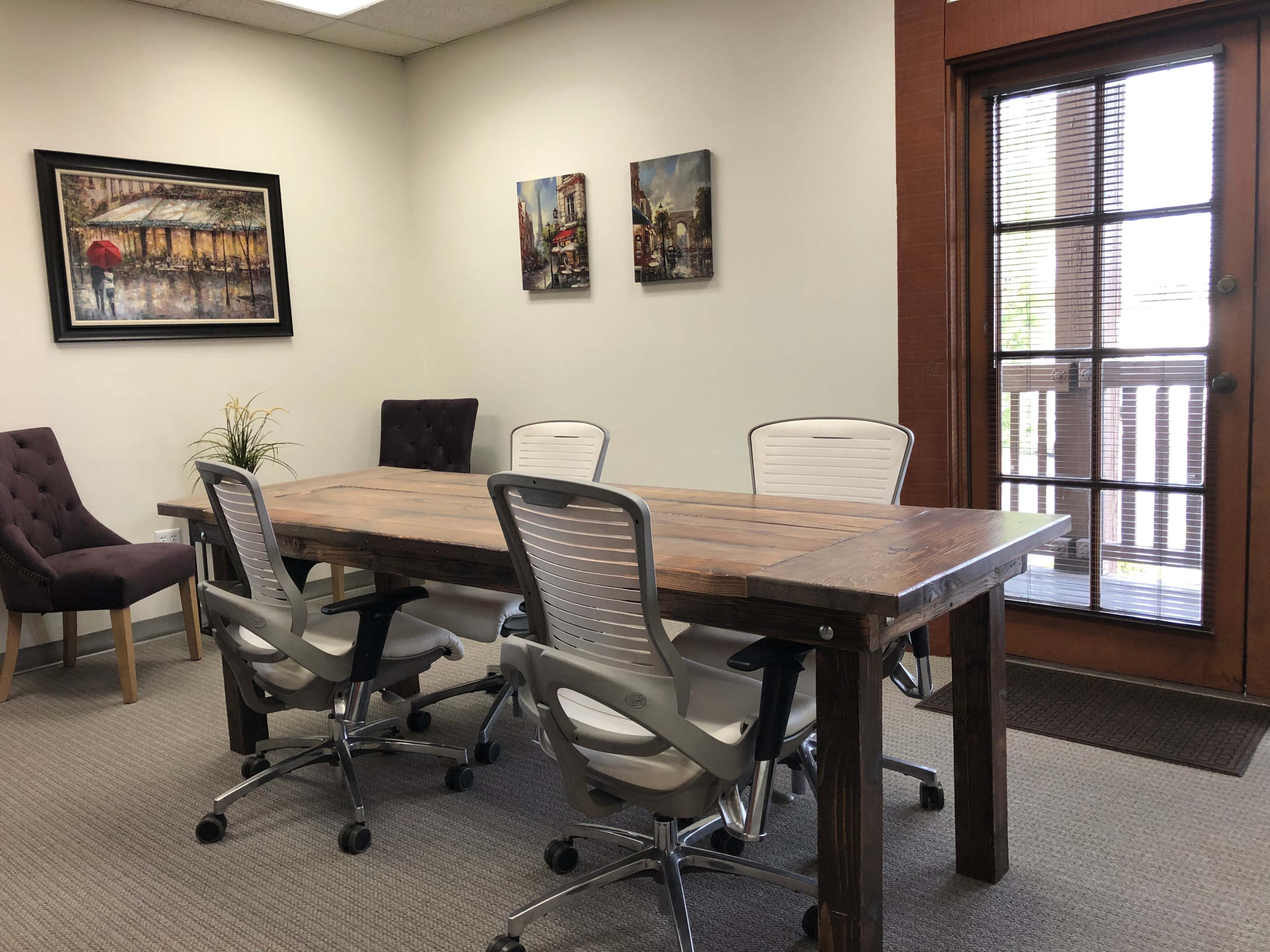 A simple conference room features a wooden table surrounded by four white office chairs, with two framed artworks on the wall and a door leading outside.