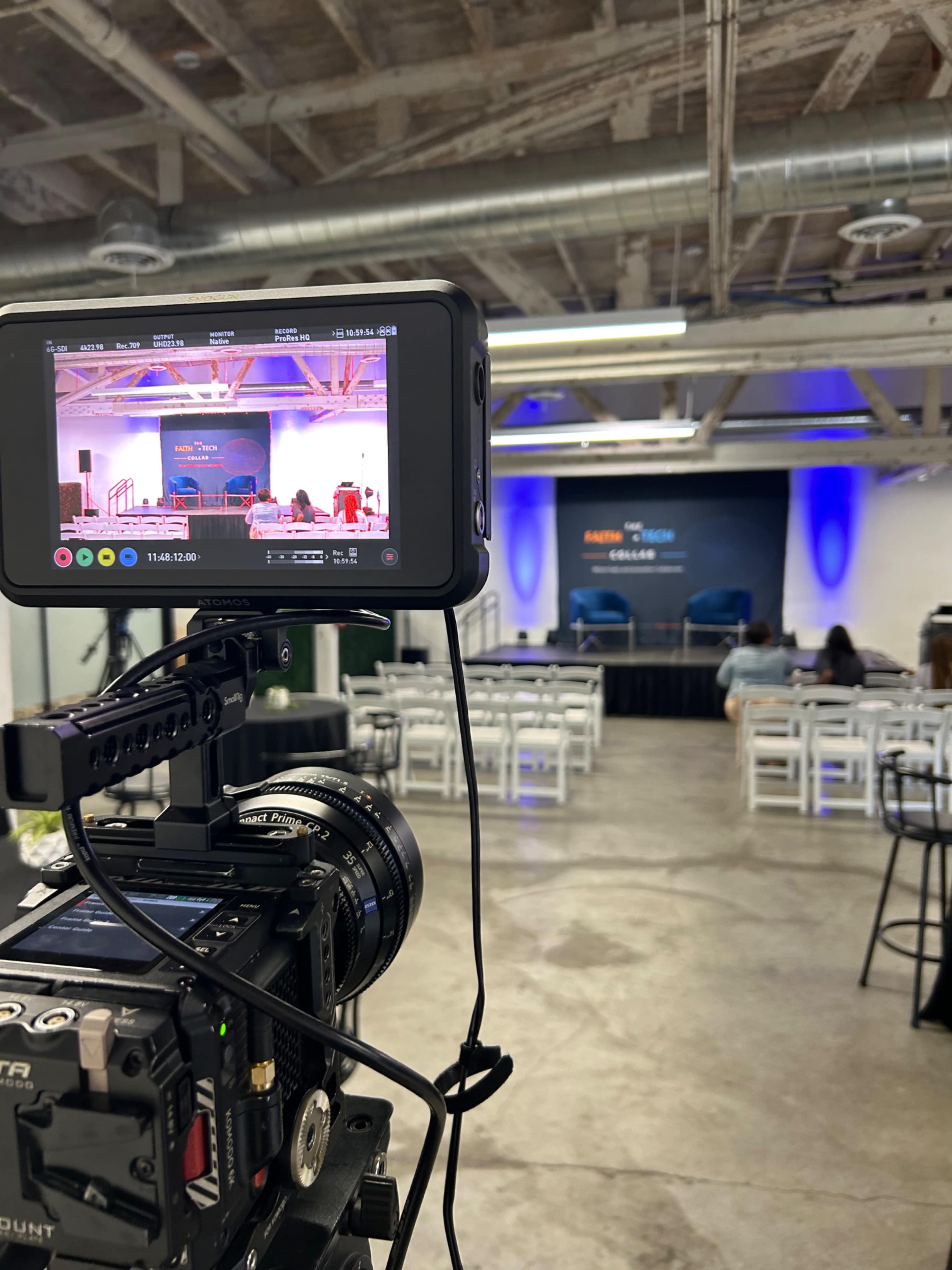 A camera mounted on a tripod captures a stage set up for an event in an industrial room with rows of empty chairs and blue lighting.