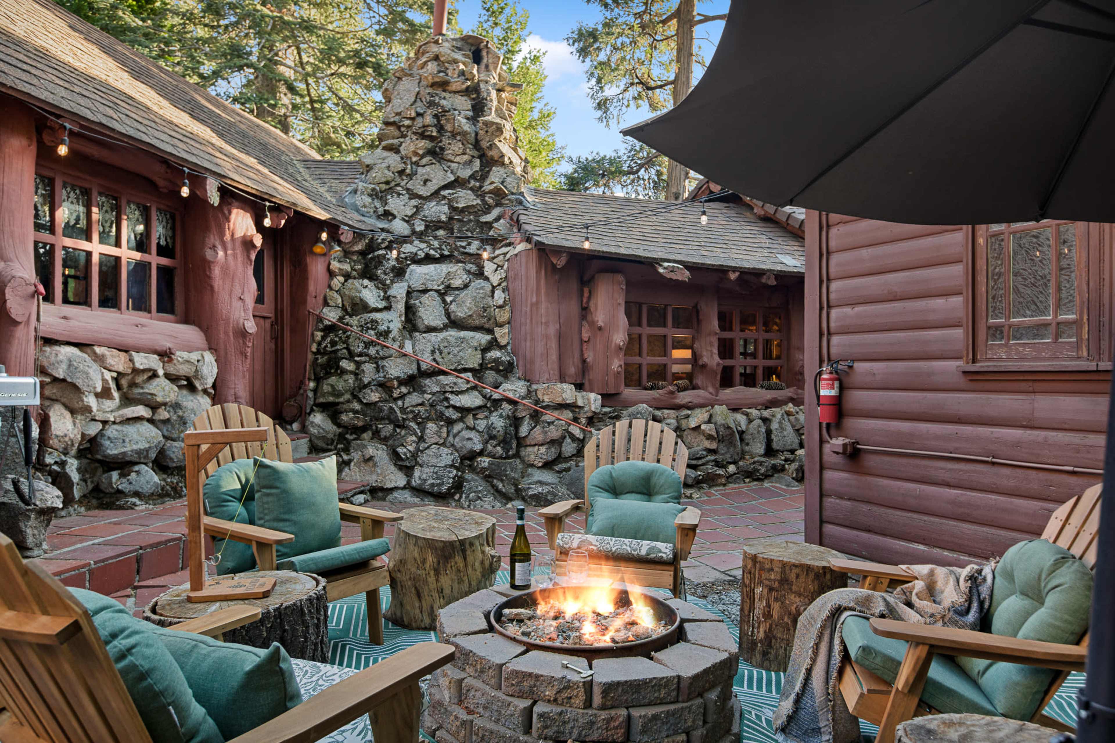 The image shows a cozy outdoor seating area with wooden chairs and a fire pit surrounded by rustic stone structures and trees.