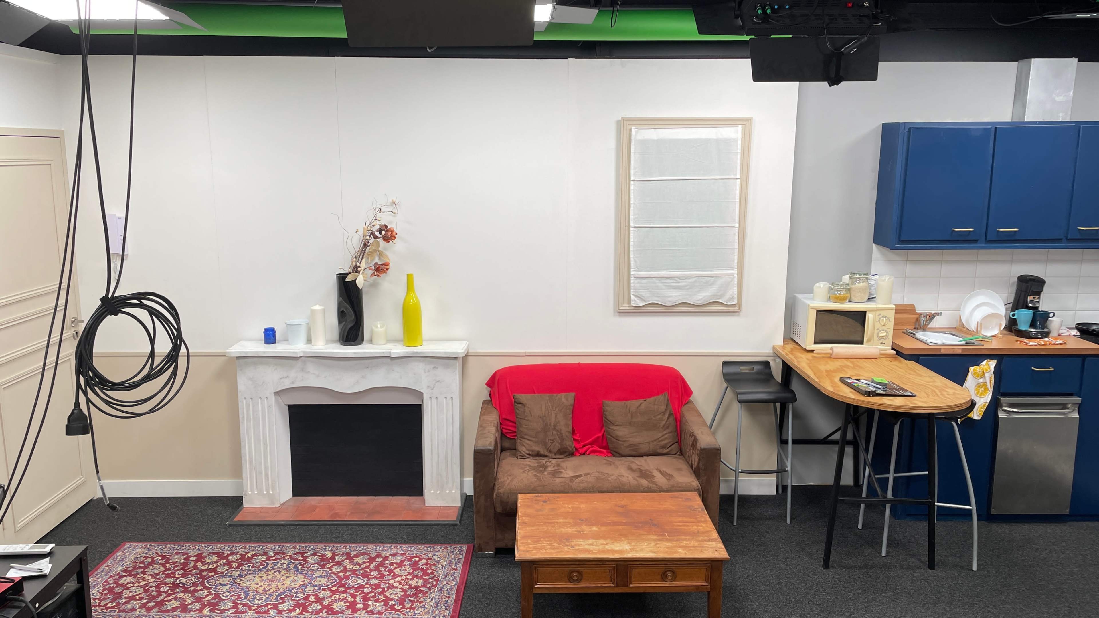 A minimalist indoor set with a brown couch, a red throw blanket, a wooden coffee table, a faux fireplace, and a kitchenette with blue cabinets and various appliances.