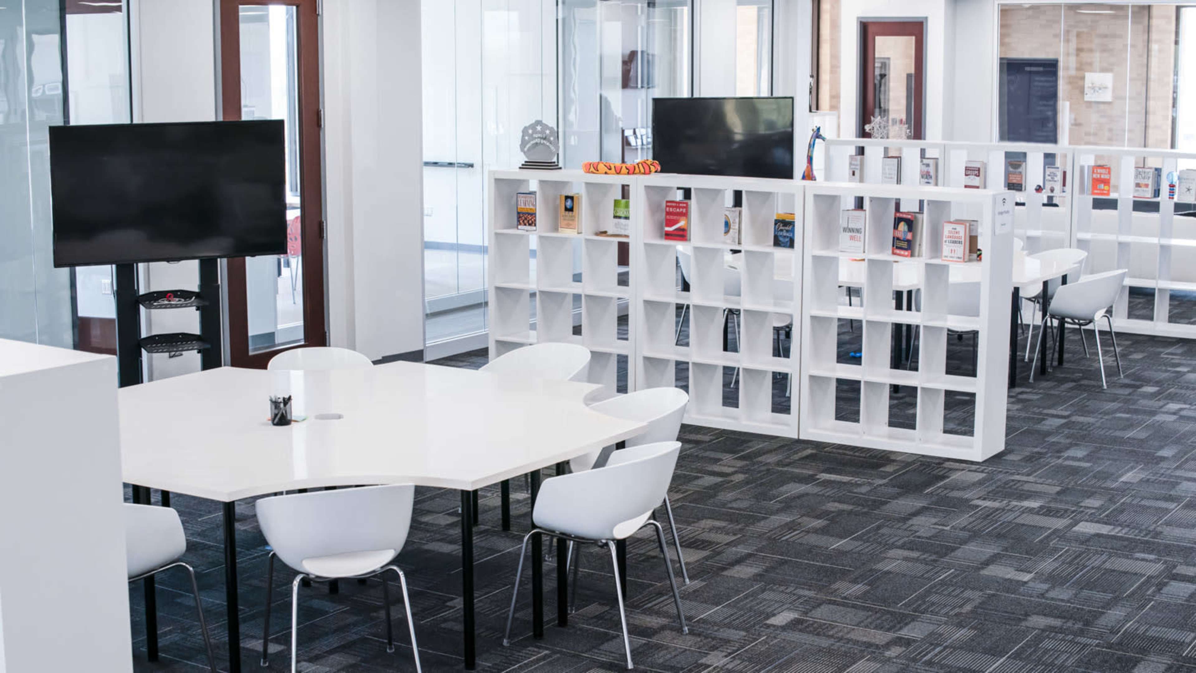 A modern office space featuring white furniture, two television screens, and a bookshelf with visible books and magazines.
