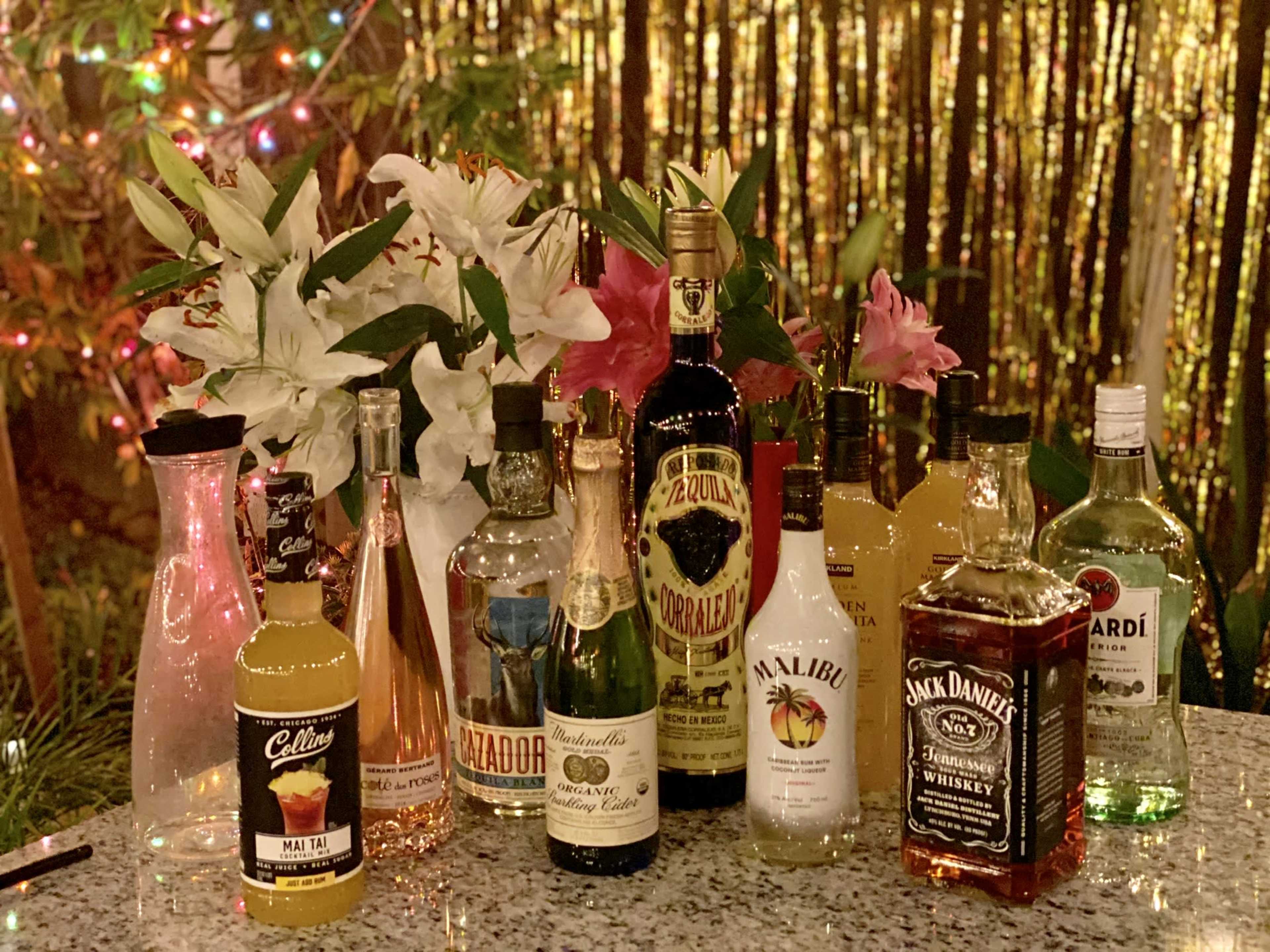 A variety of alcoholic drinks arranged on a table alongside decorative flowers, with a festive backdrop of lights.
