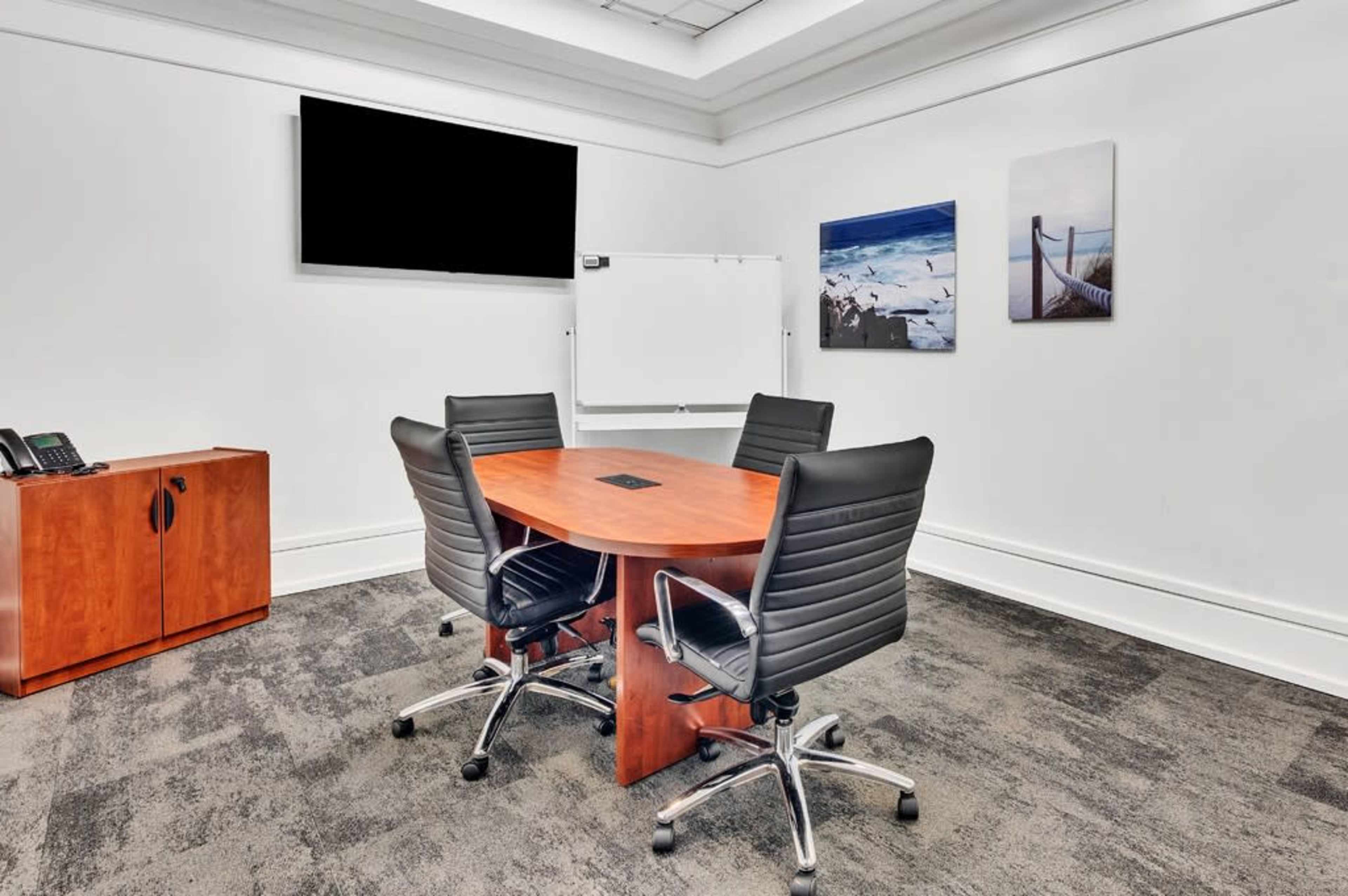 The image shows a small conference room featuring a round wooden table surrounded by four black swivel chairs, with a whiteboard and wall-mounted television in the background.