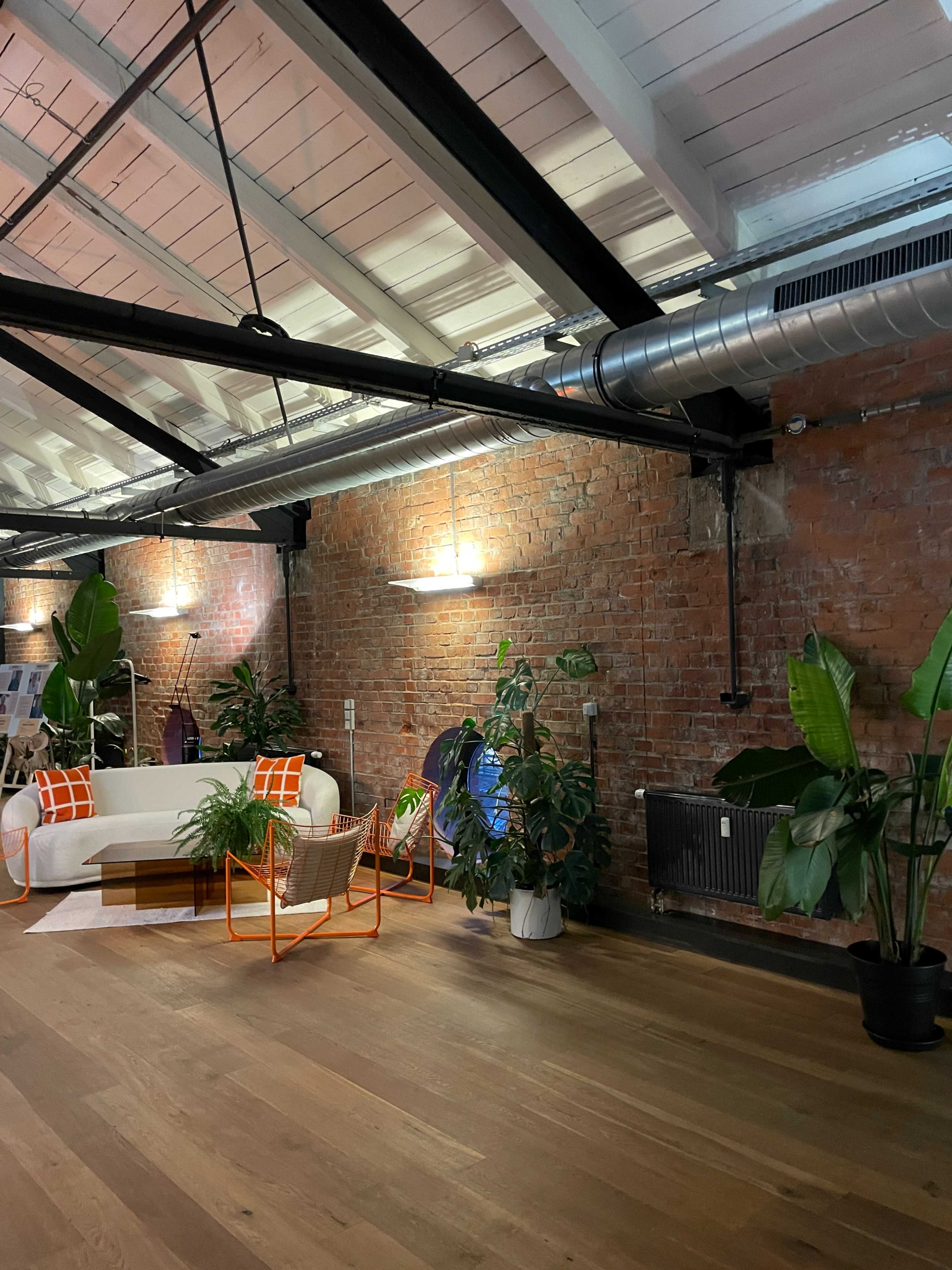 The image shows a spacious, modern interior with exposed brick walls, a white sofa and colorful seating, along with various plants and wooden flooring.