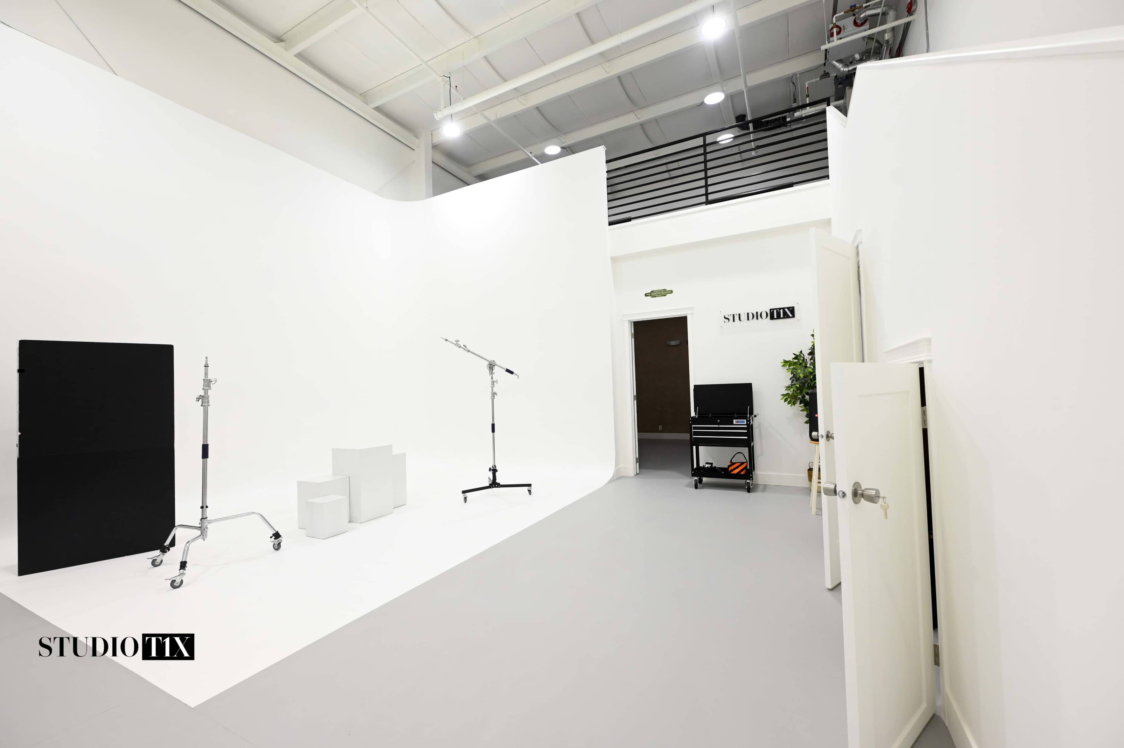 The image shows a spacious, well-lit studio with white walls, various equipment stands, and a door leading to another room.