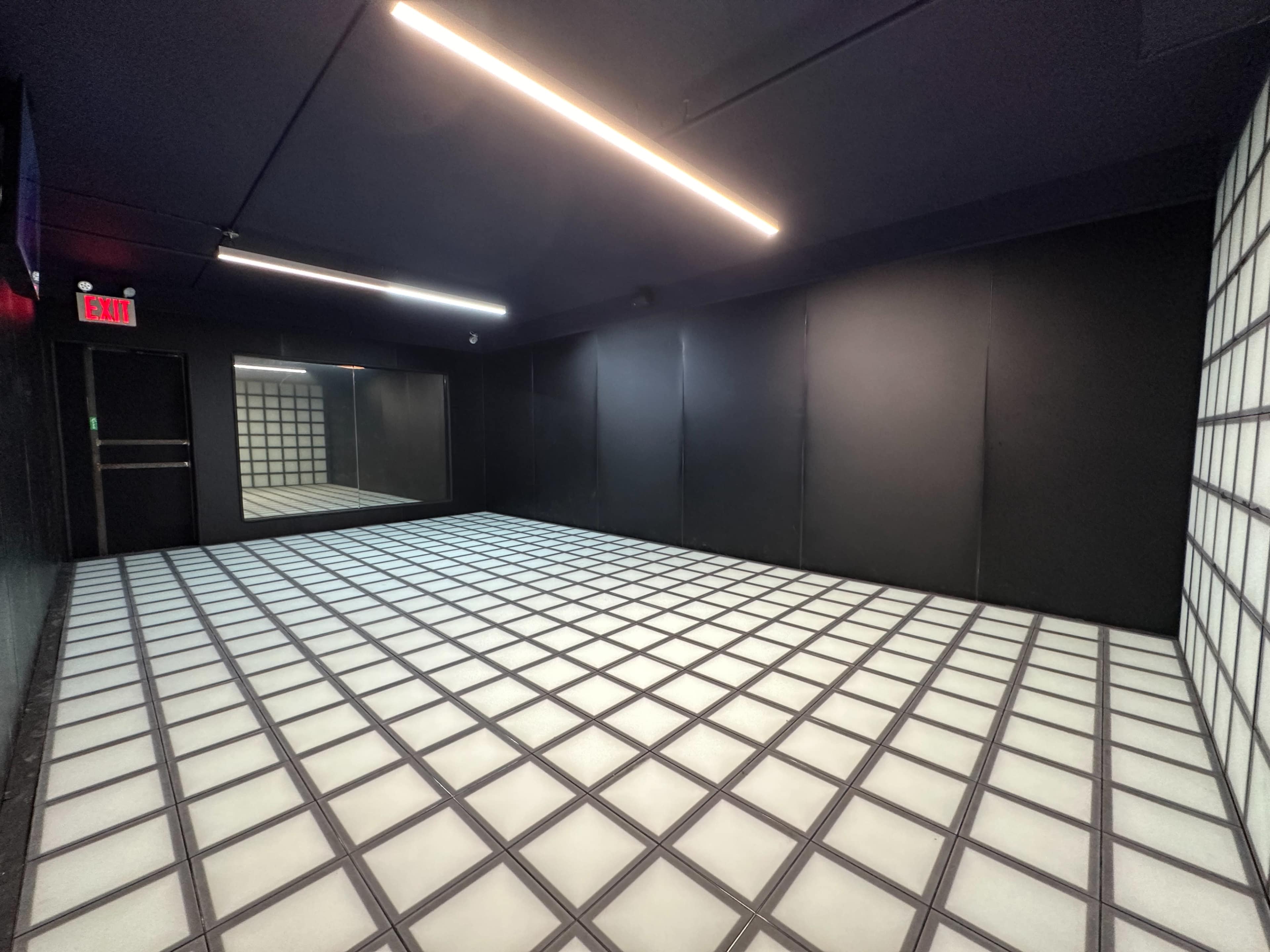 A minimalist room with a black ceiling and walls, featuring a grid-patterned floor in white and gray tiles, and a mirror on one wall.