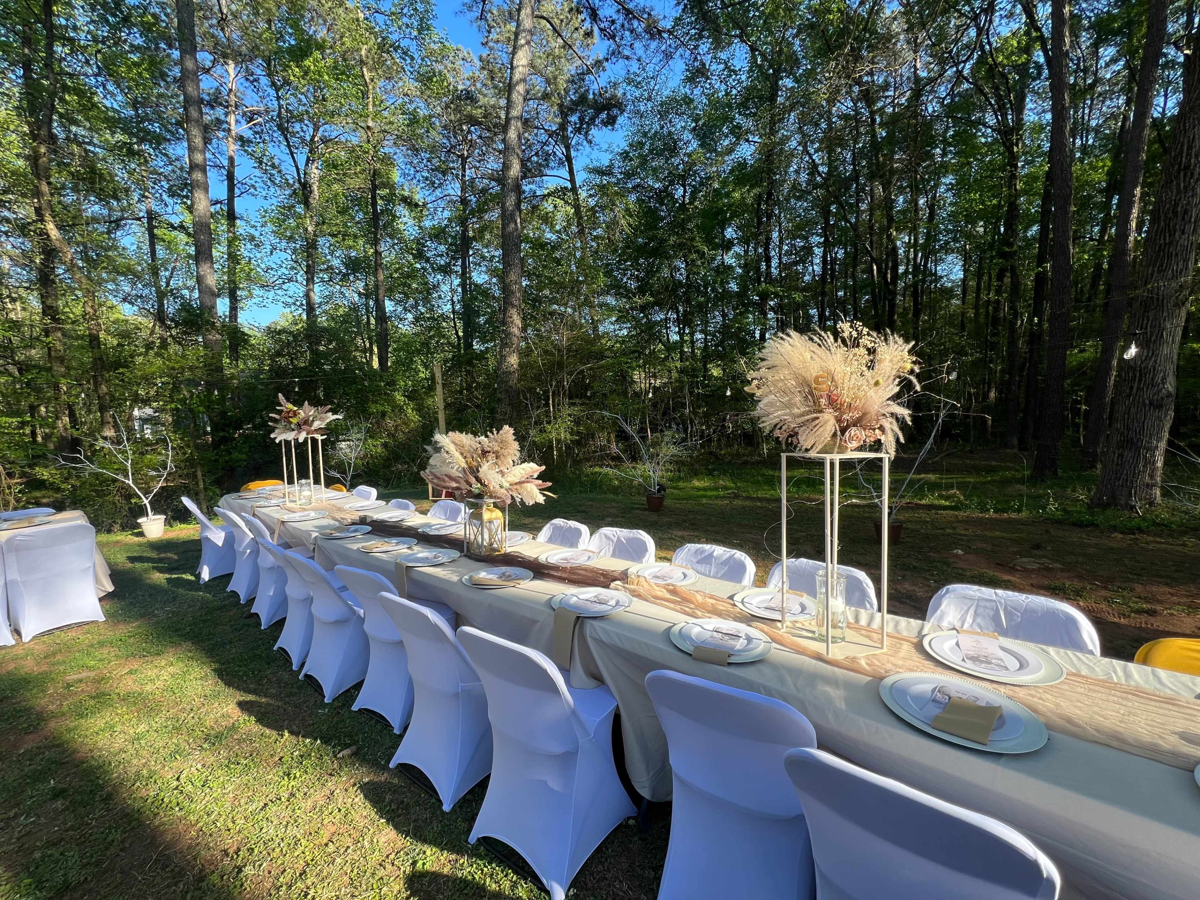 A long outdoor dining table is set with decor and chairs amidst a wooded area.