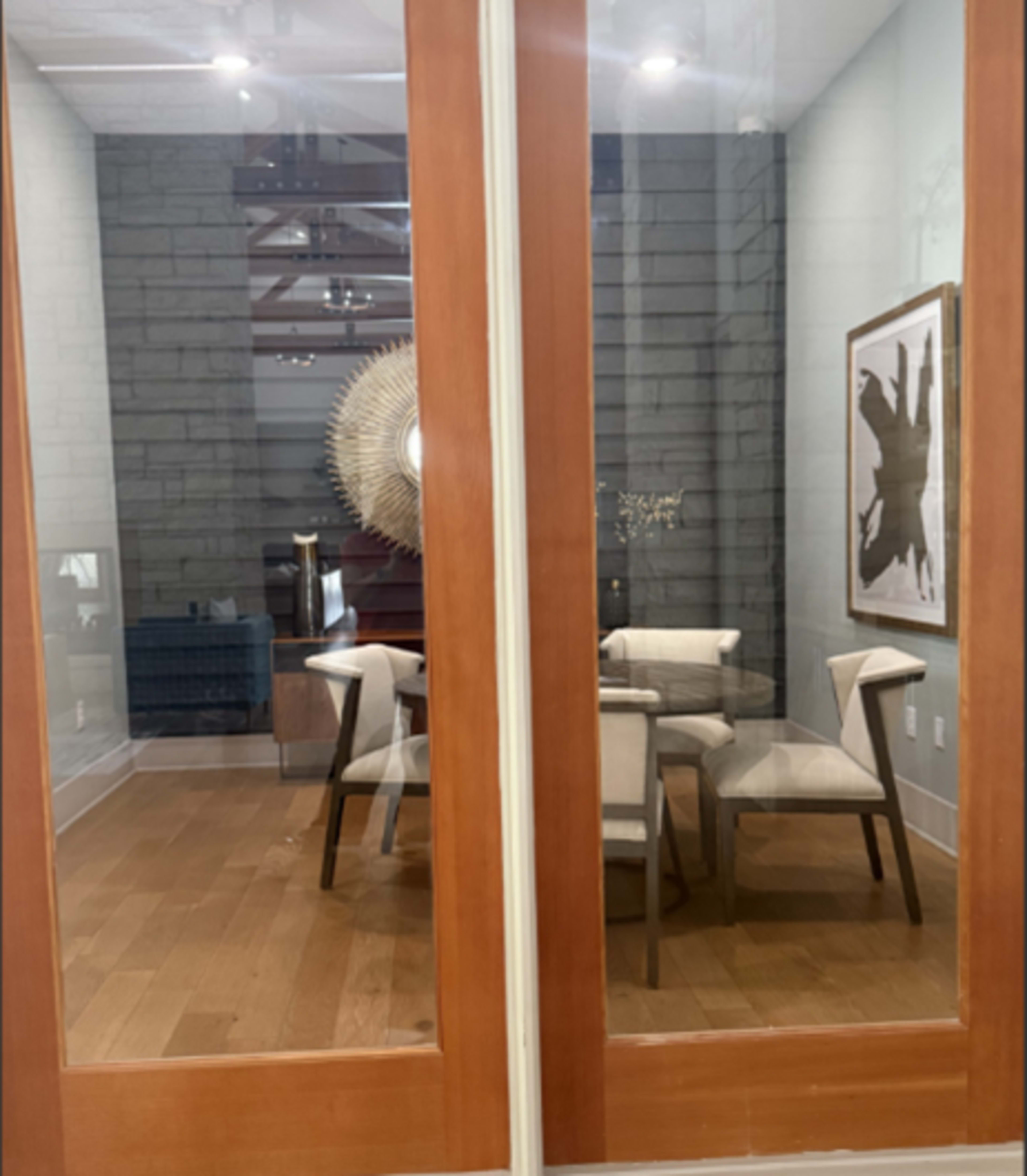 The image shows a modern room with wooden flooring and a glass wall, featuring a round table and several chairs, along with a large decorative mirror.