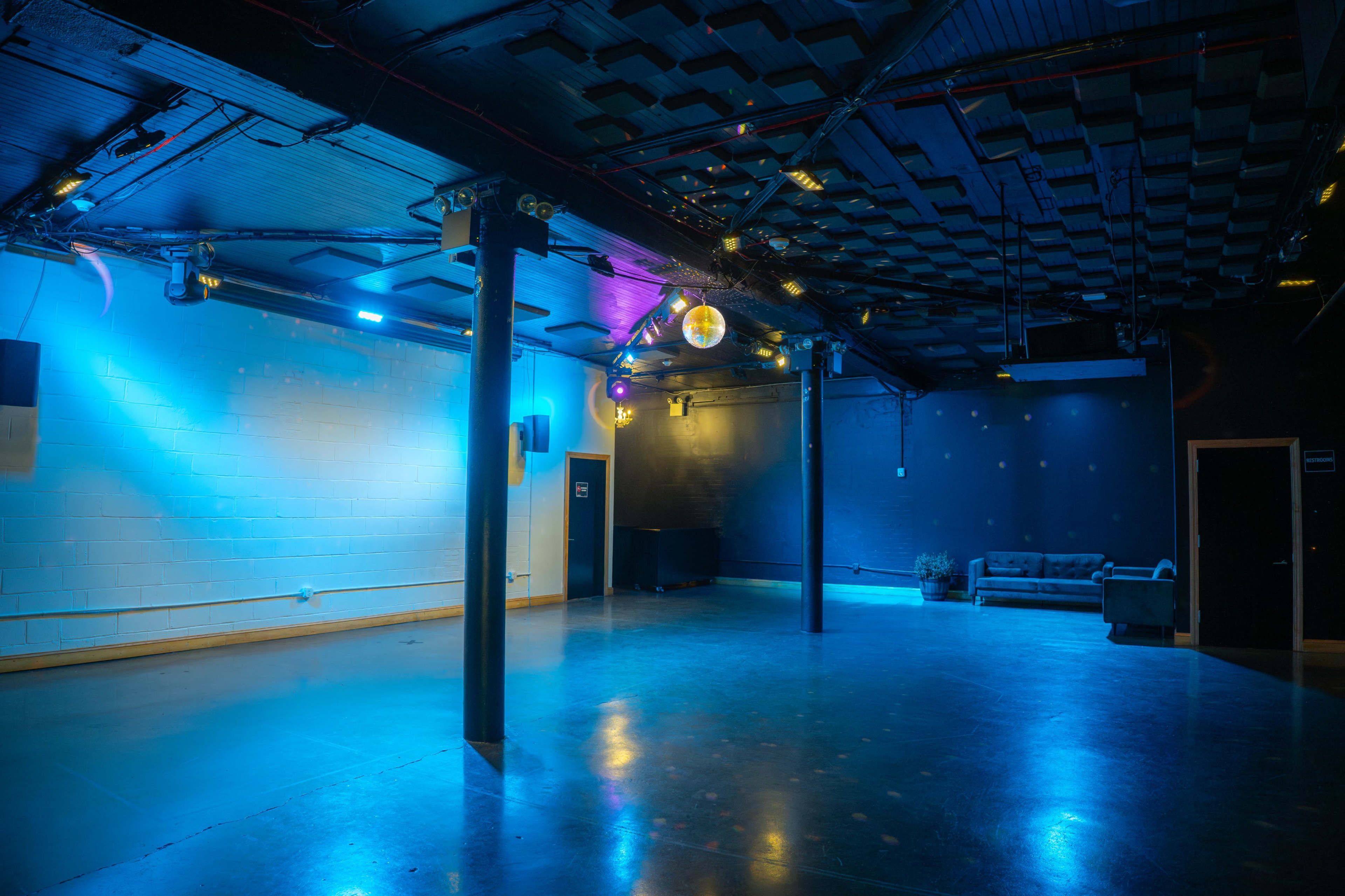 The image shows an empty event space with blue lighting, concrete floors, and a couch against one wall.