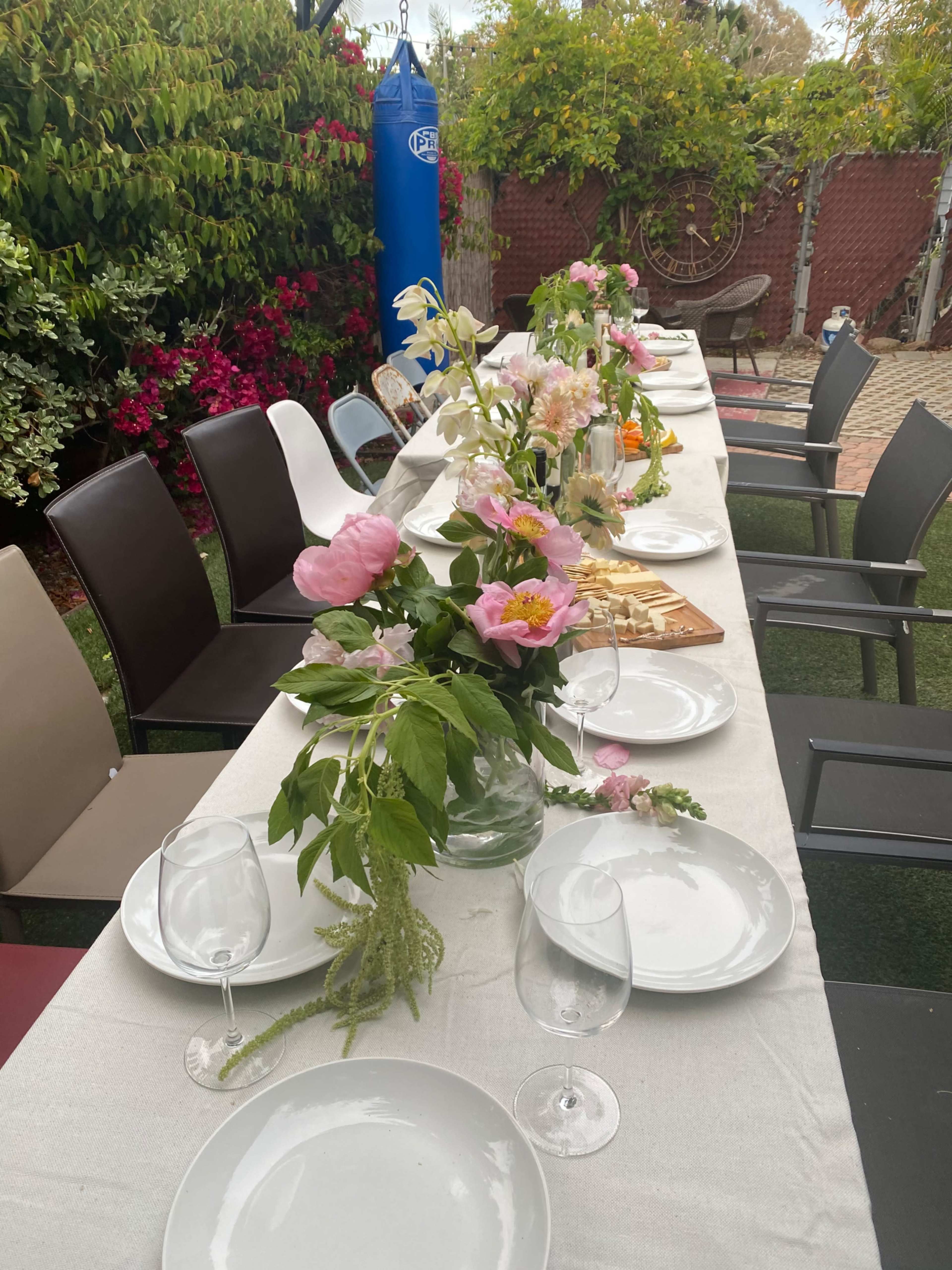 A long dining table is set outdoors with white plates and glassware, decorated with flower arrangements, and surrounded by chairs under a lush garden backdrop.