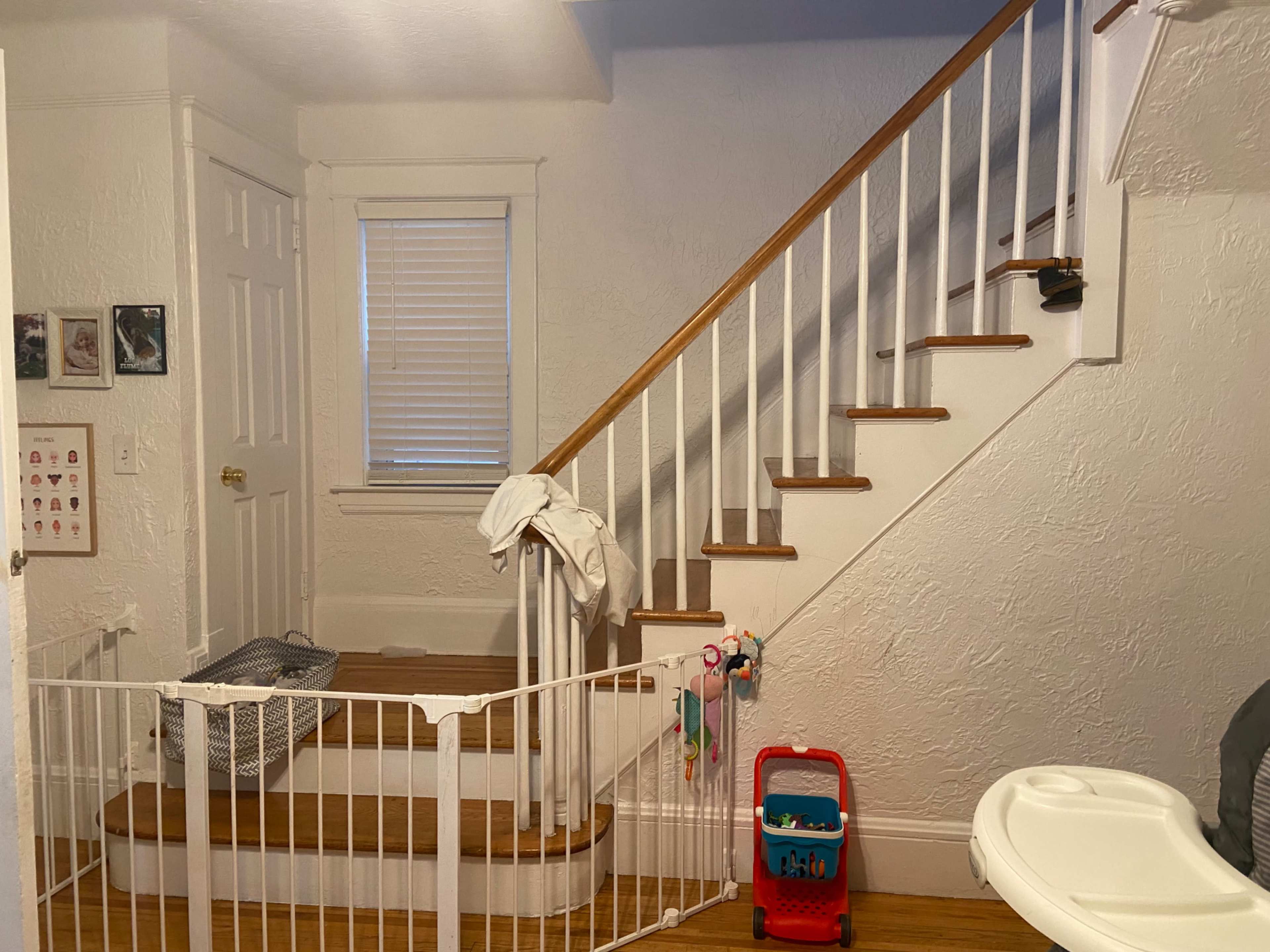 The image shows an interior entryway with a staircase, a baby gate, and a small play area featuring a toy cart and a high chair.