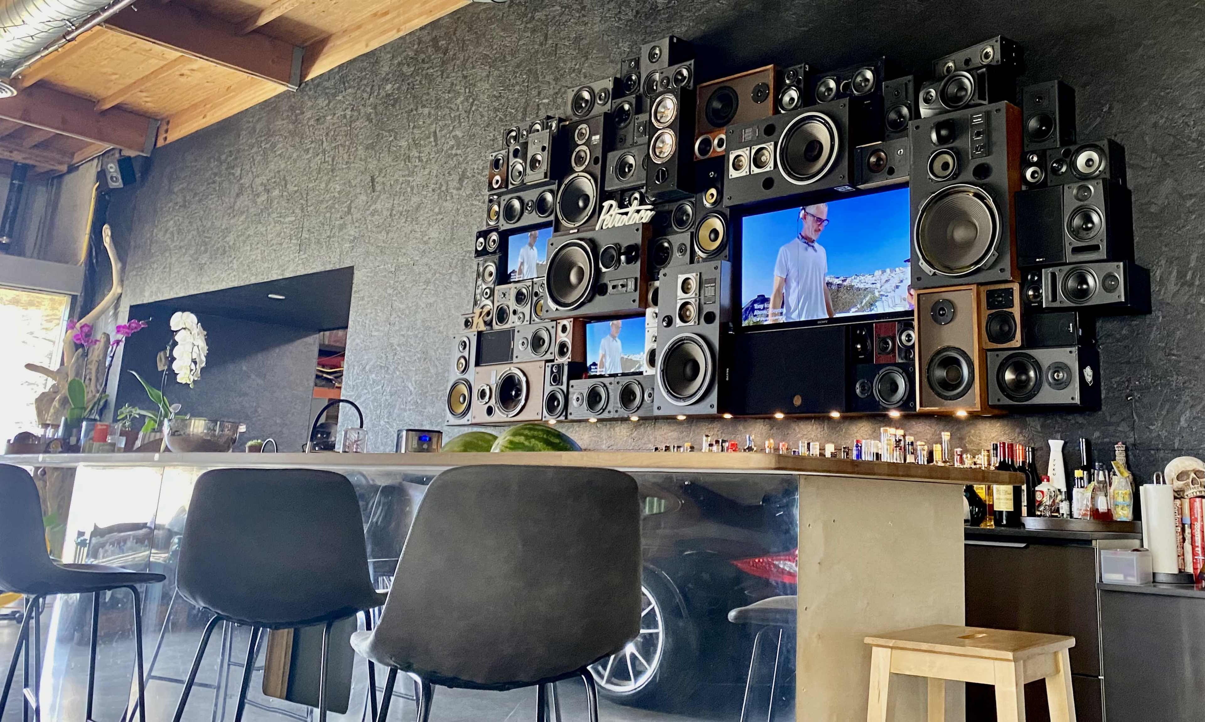 The image shows a wall-mounted arrangement of speakers and televisions in a modern bar area with a concrete counter and high stools.