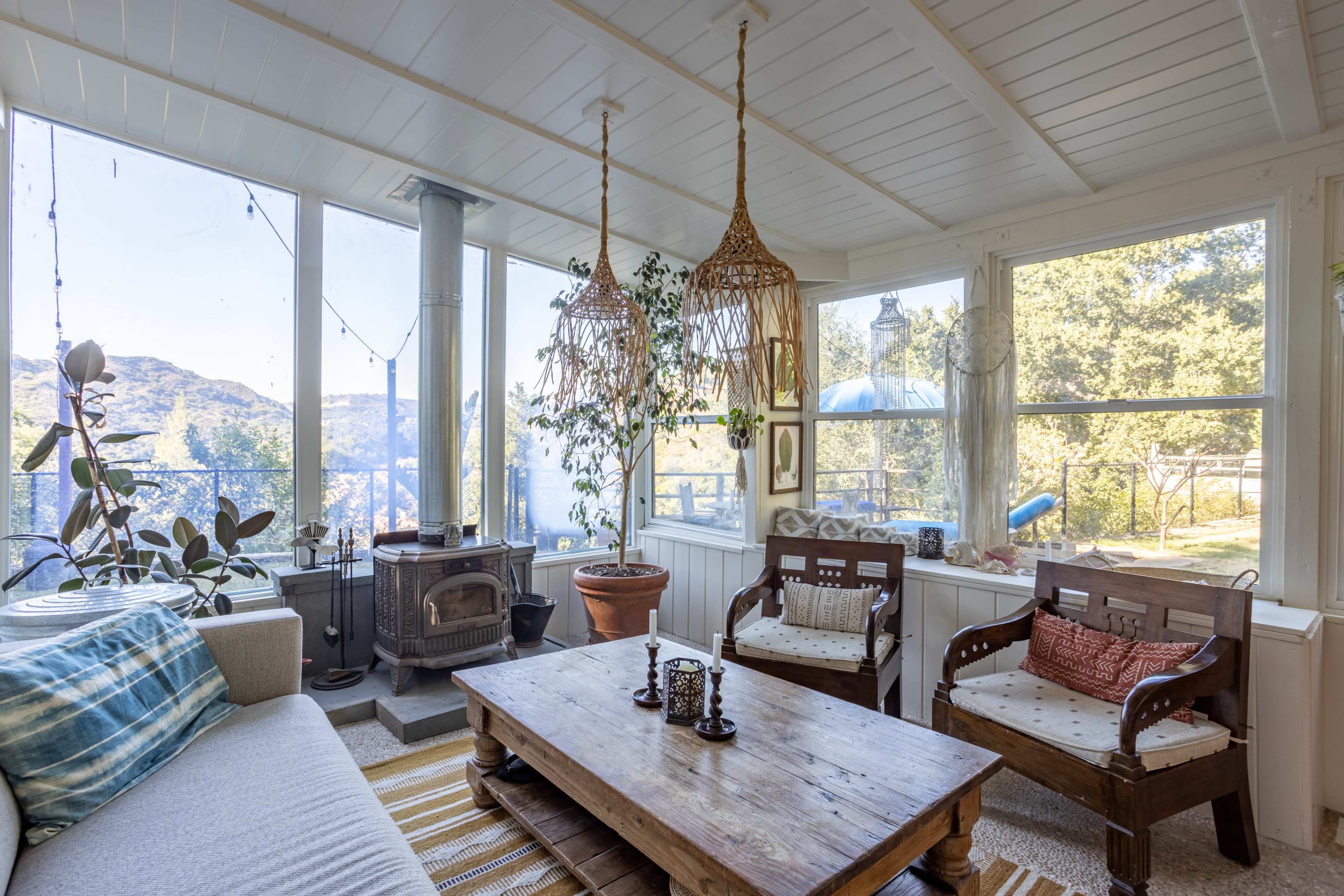 A sunlit sunroom with a cozy seating area, a wooden table, and large windows offering views of hills and trees outside.