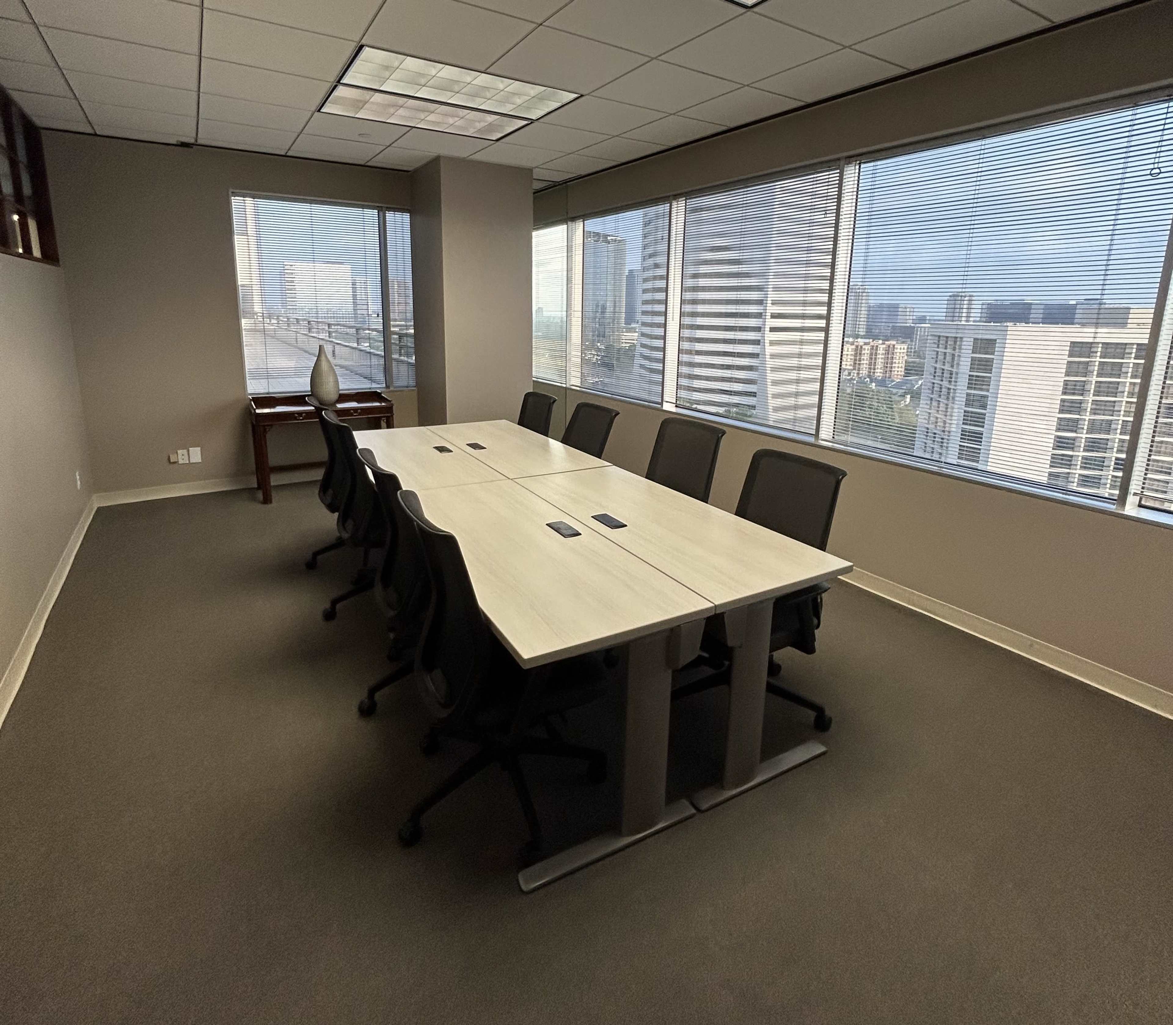 The image shows a modern conference room with a long table and several black chairs, overlooking a cityscape through large windows.