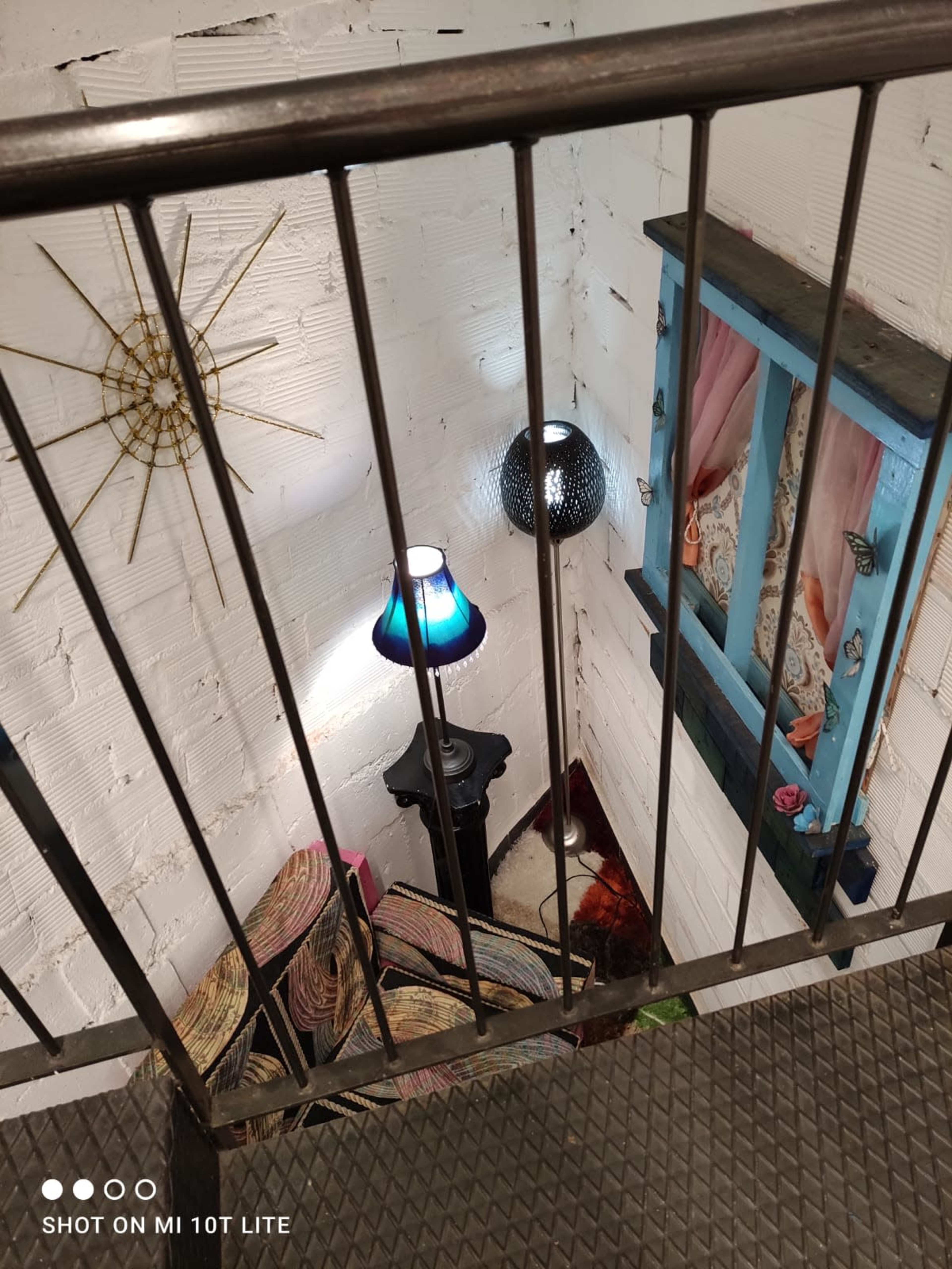 The image shows a staircase overlooking a cozy corner with a patterned rug, a lamp, and a decorative window framed in blue.