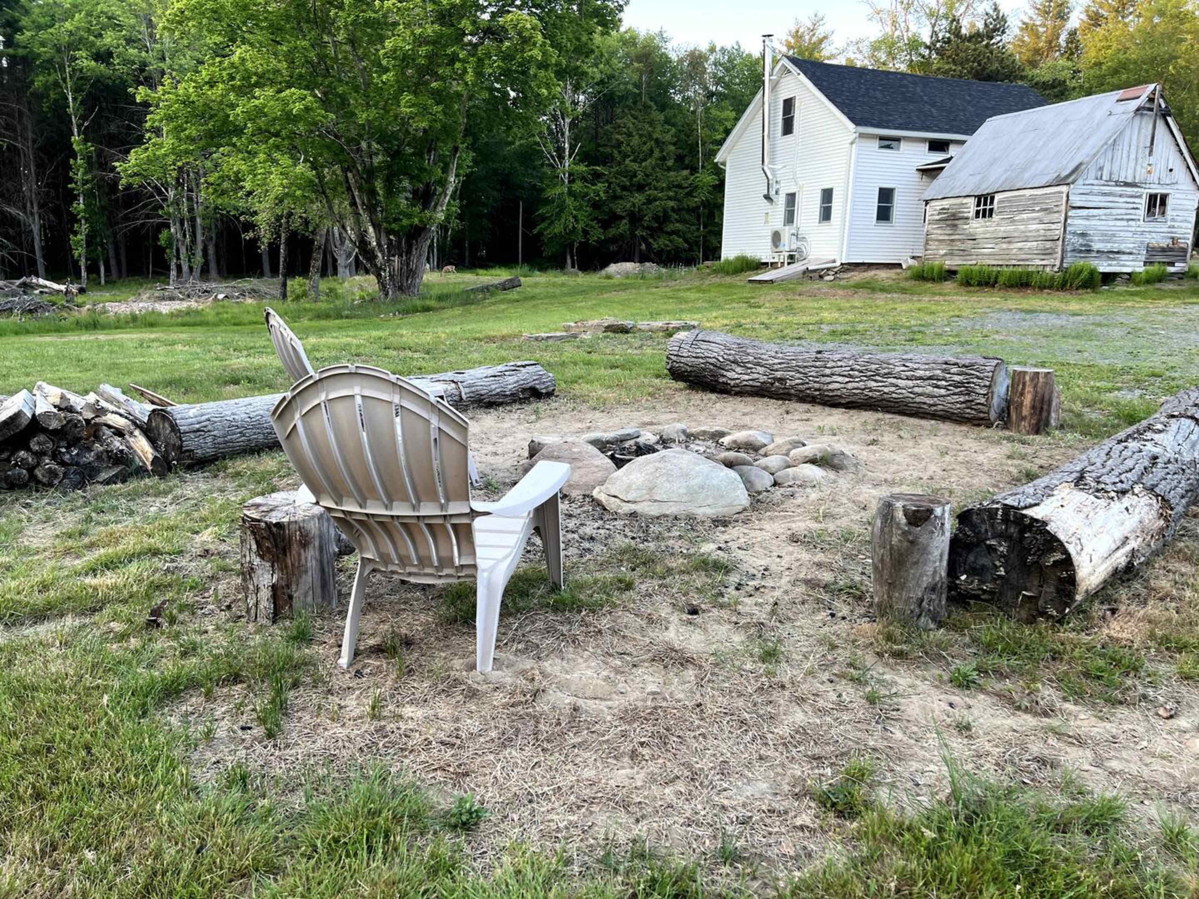 A single plastic chair sits beside a fire pit made of stones, surrounded by logs and a grassy area next to a weathered house in a forested setting.