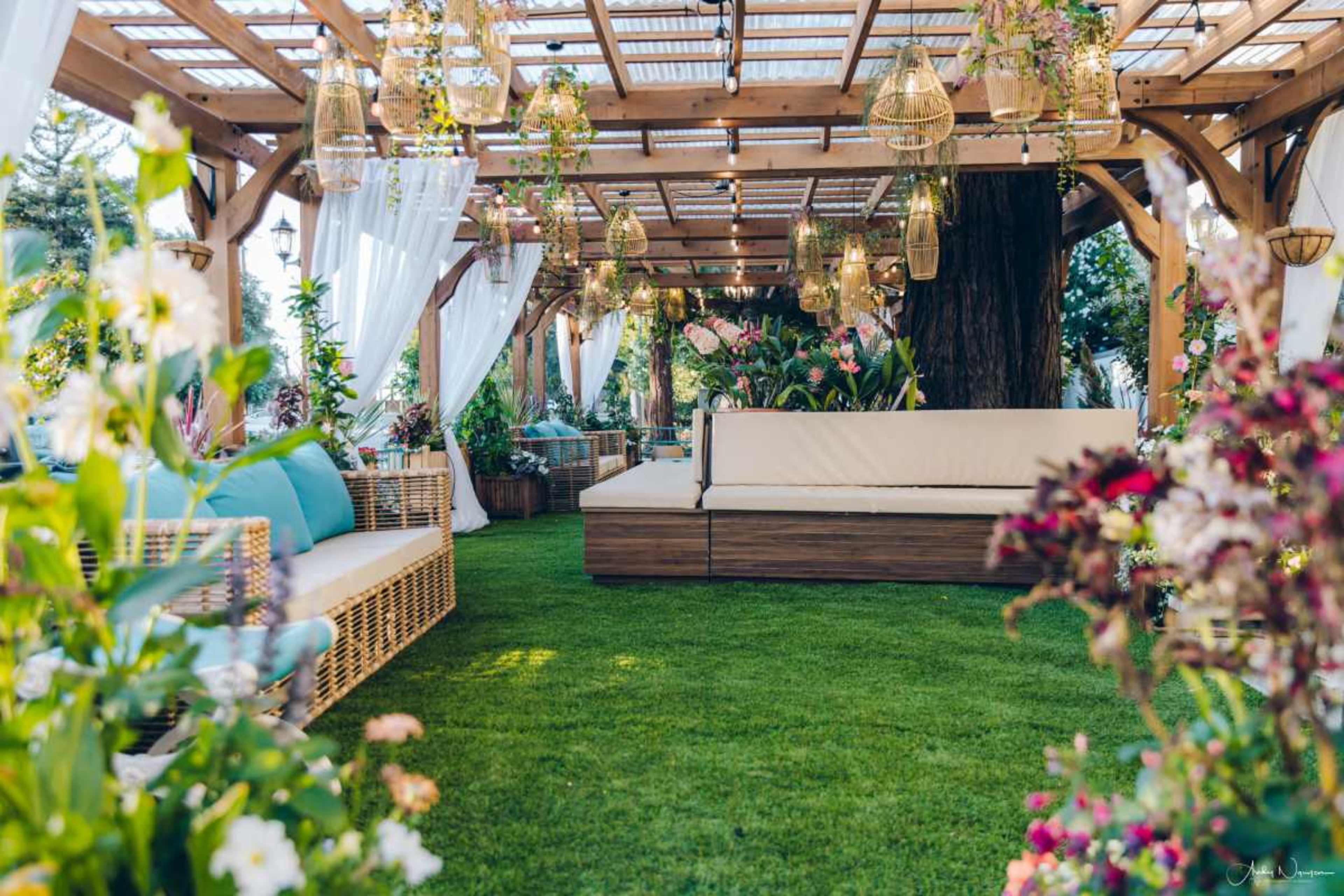 The image depicts a cozy outdoor seating area with wooden pergolas, surrounded by lush greenery and decorative plants.