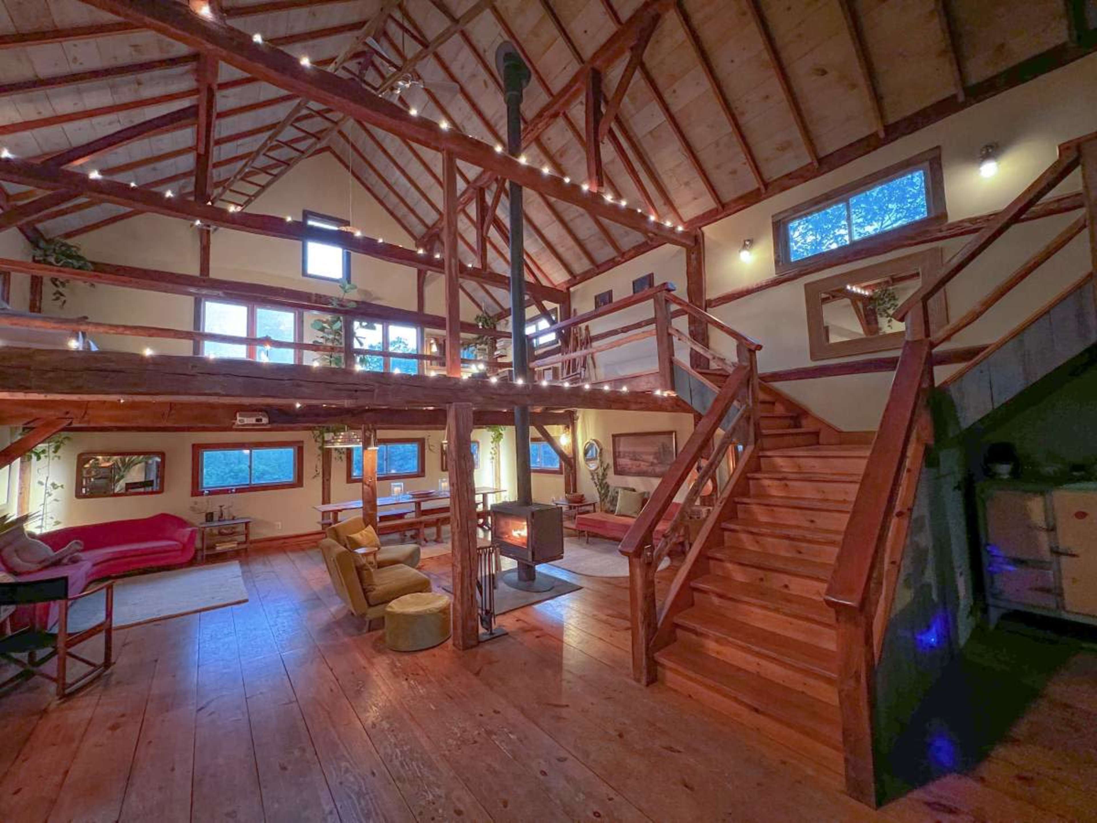 The image shows a spacious wooden interior of a cabin-style home featuring a staircase, cozy seating areas, and large windows letting in natural light.
