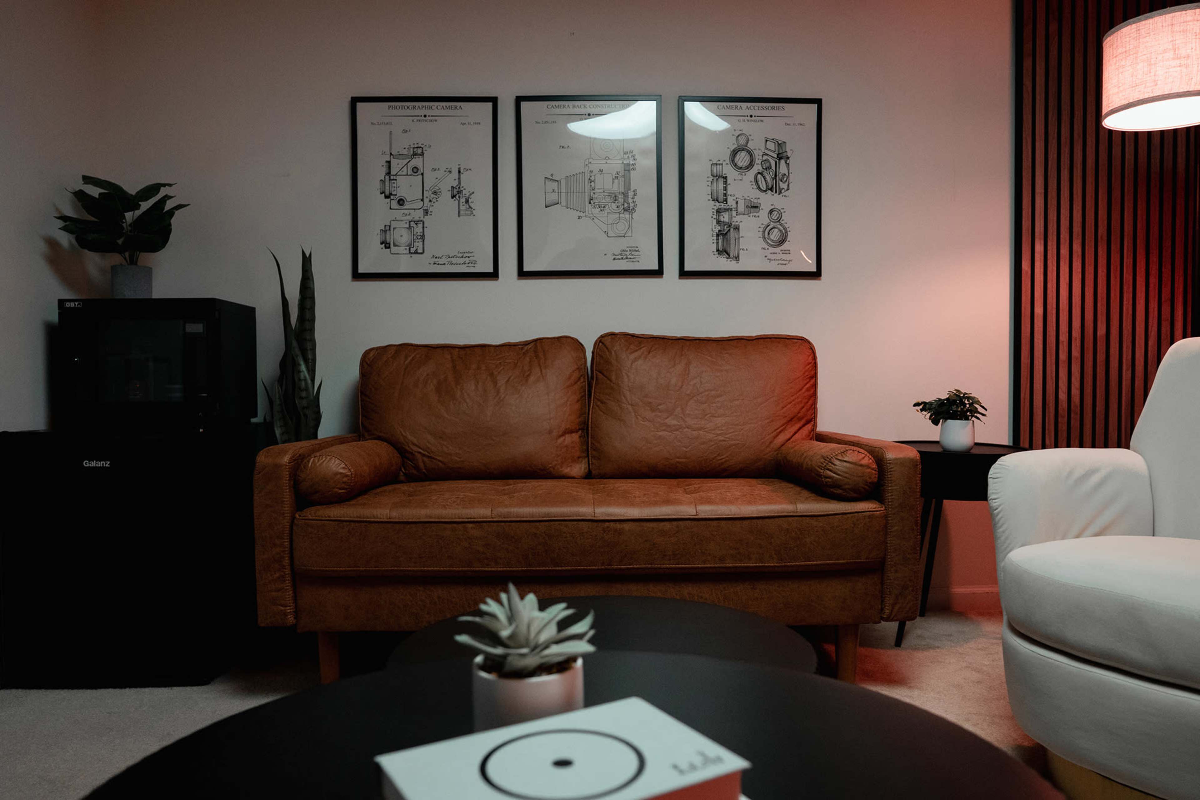 A living room with a brown leather couch, two framed drawings on the wall, a small black refrigerator, and a white armchair.