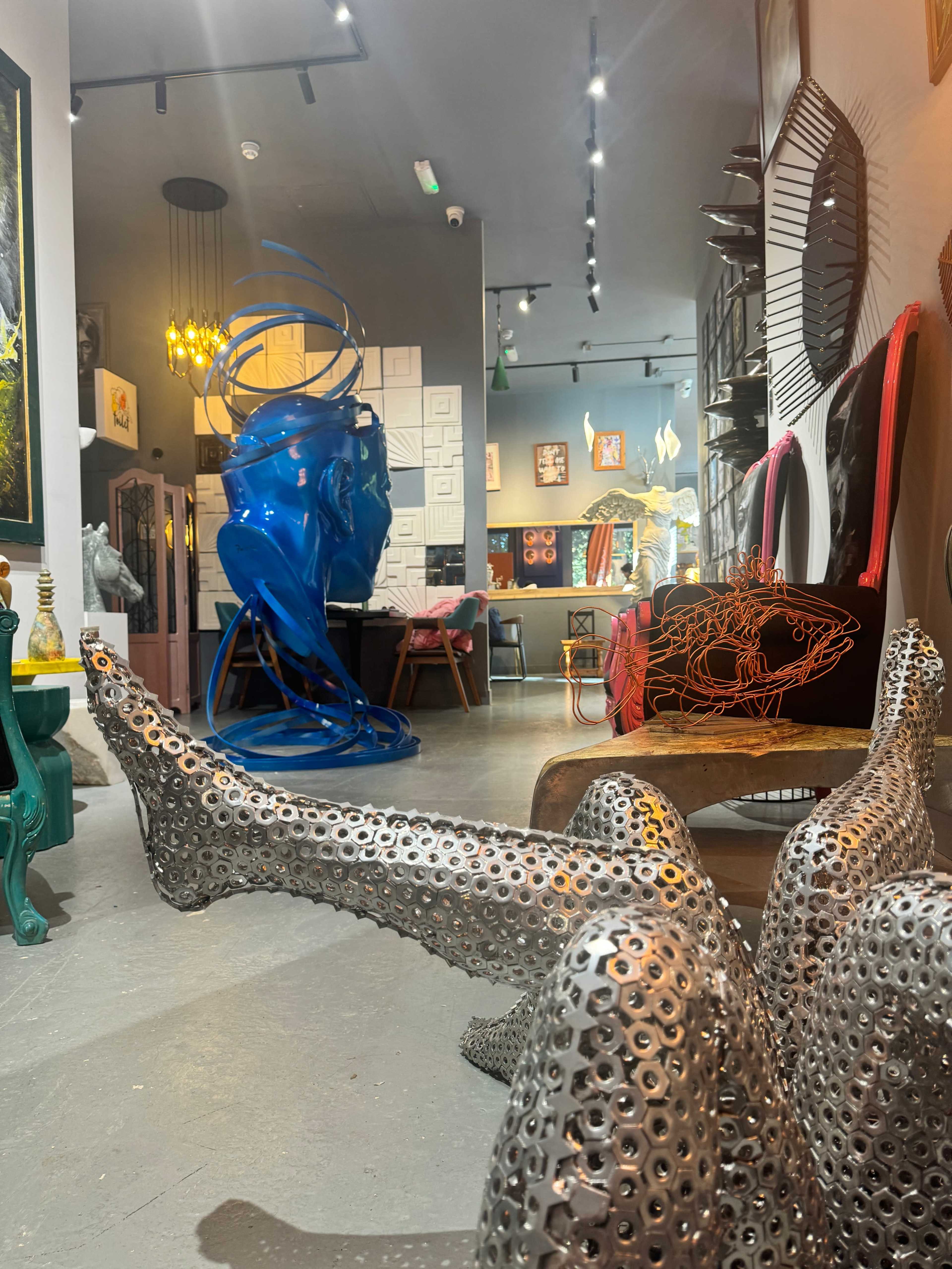 The image features an art gallery with a metallic sculpture resembling human legs in the foreground, colorful artwork on the walls, and various modern furnishings in the background.