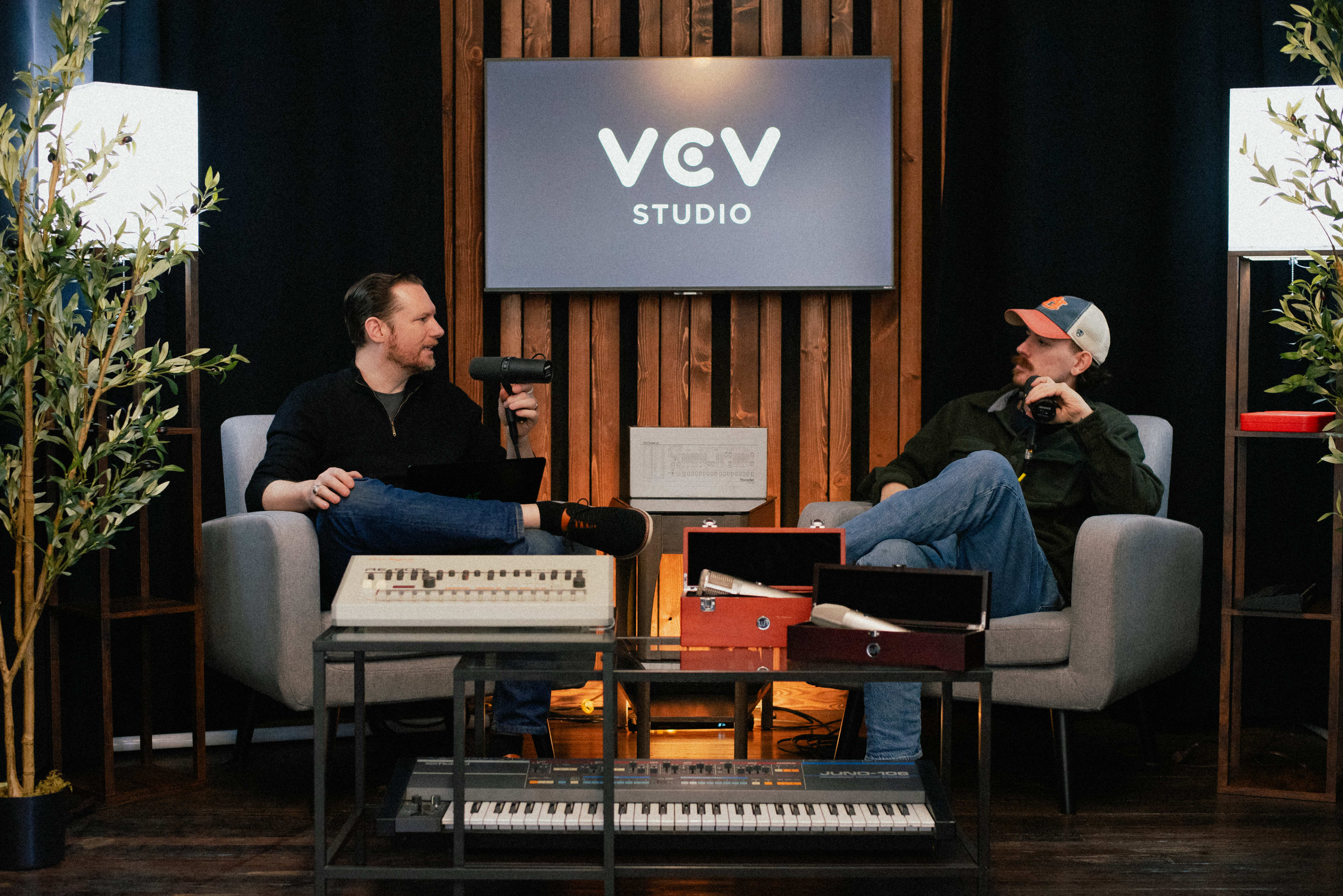 Two men are seated in a cozy studio setting, engaged in conversation, with musical instruments and production equipment placed in front of them.
