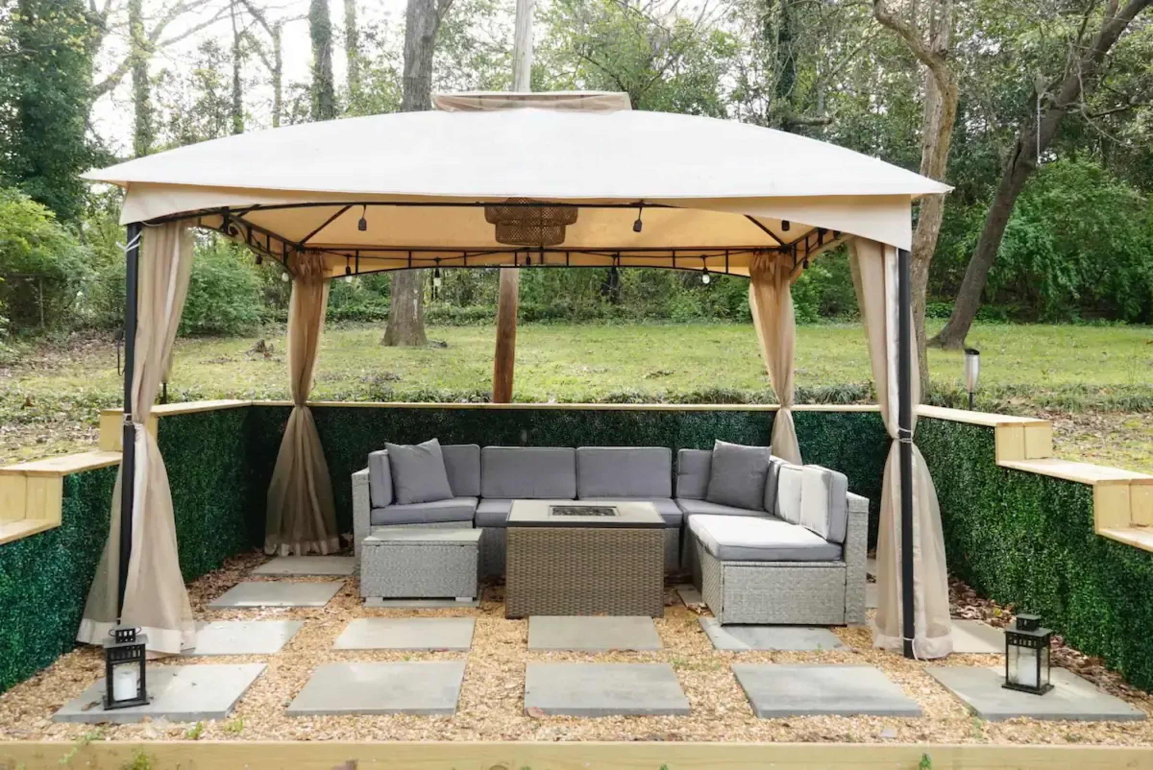 A gazebo with a sectional seating area and fire pit is situated on a gravel patio surrounded by greenery.