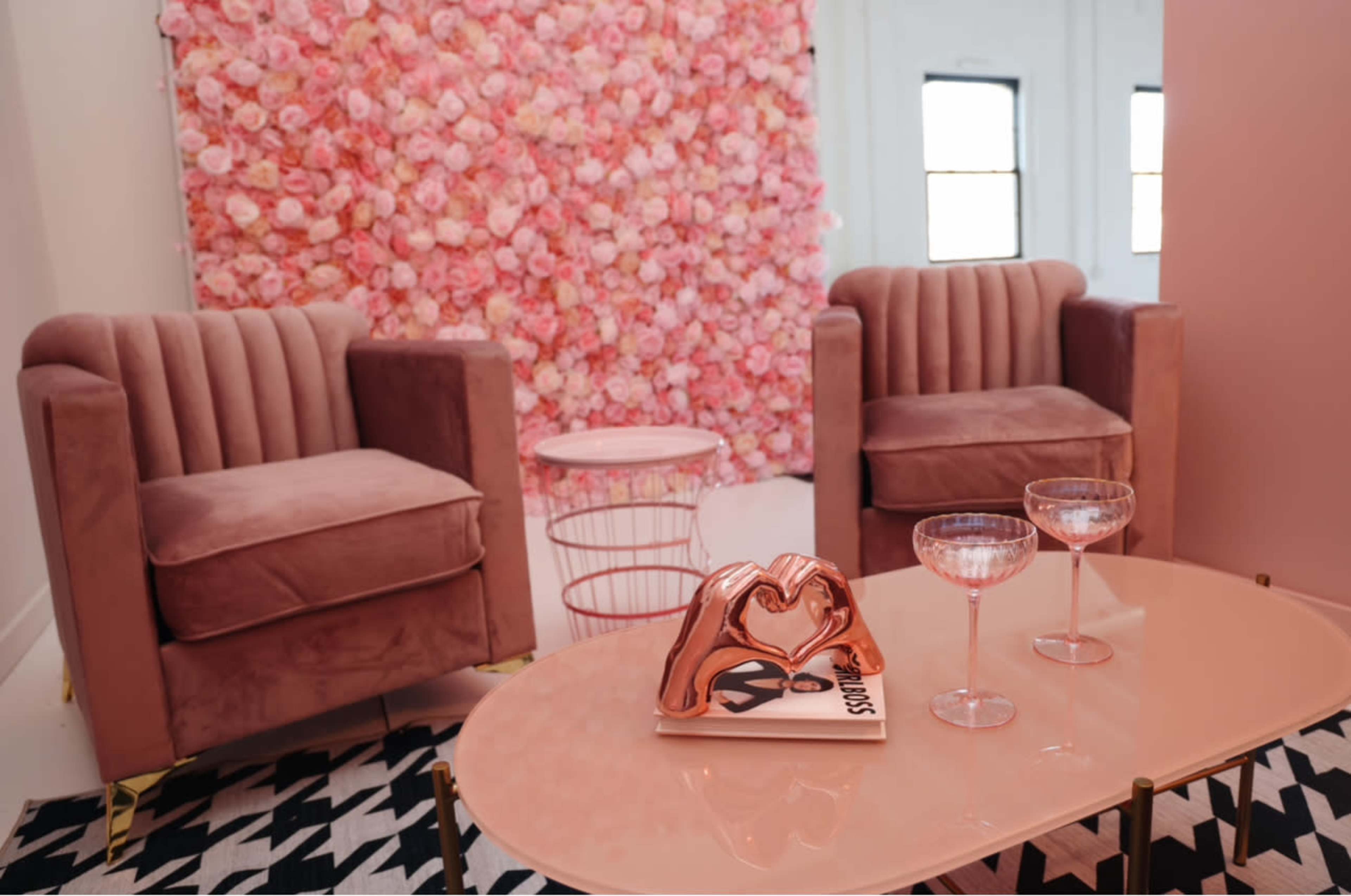 A room features two pink velvet chairs facing a small table, with a floral backdrop of pink flowers behind them.