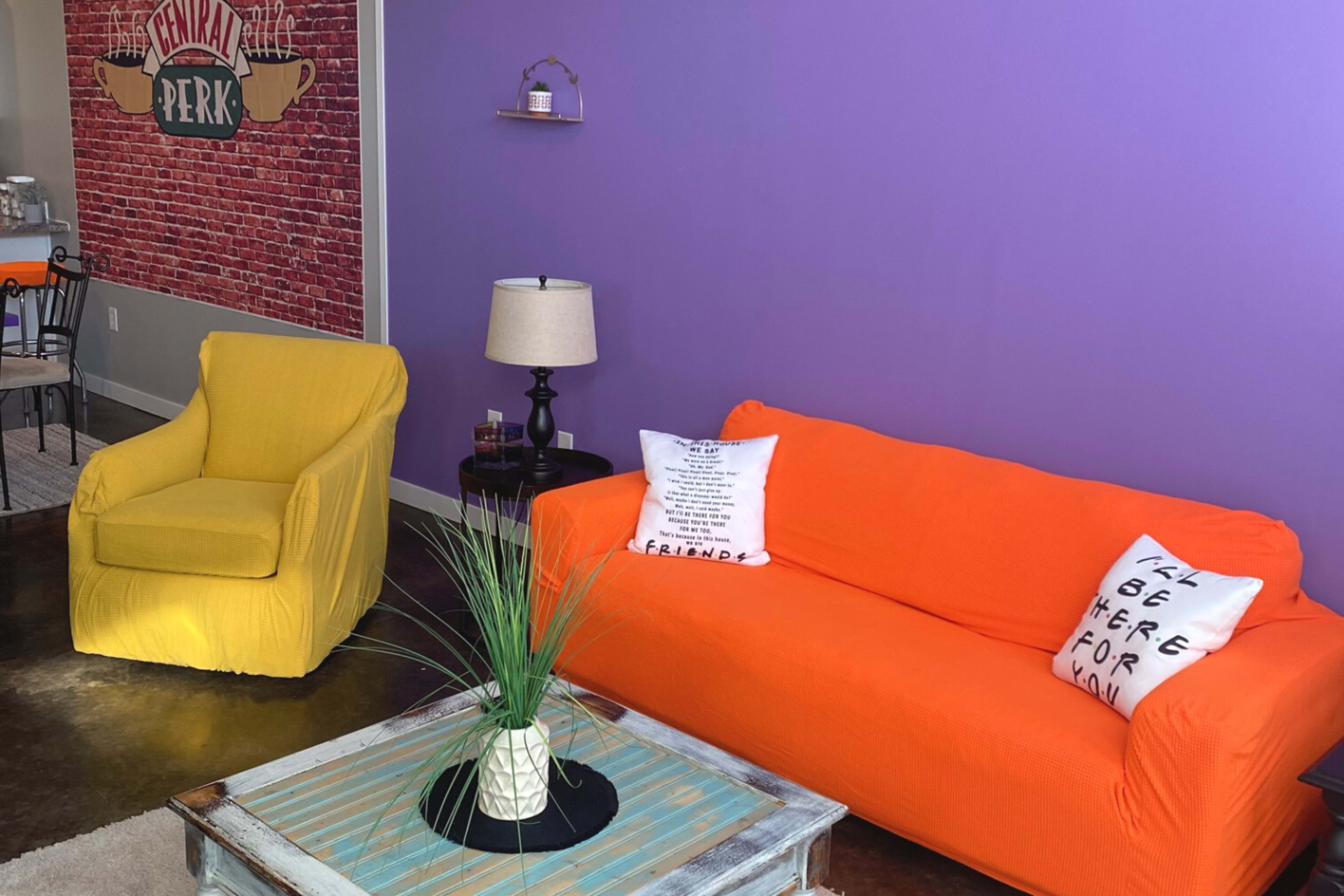 A cozy living room setup featuring an orange sofa, a yellow armchair, and a coffee table adorned with a small plant, against a purple wall.