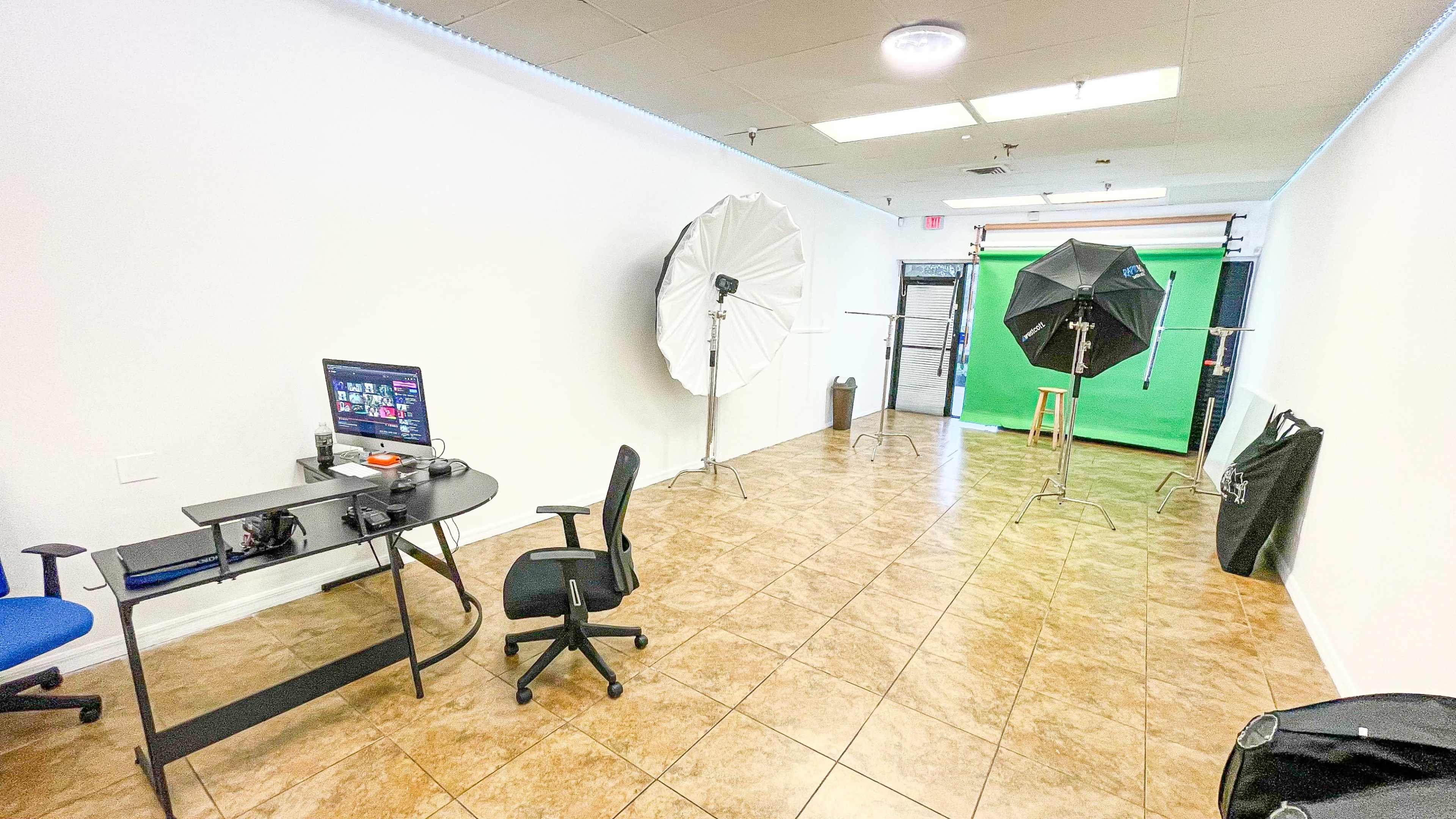 A studio space with a green screen, two lighting softboxes, a computer desk with an office chair, and tiled flooring.