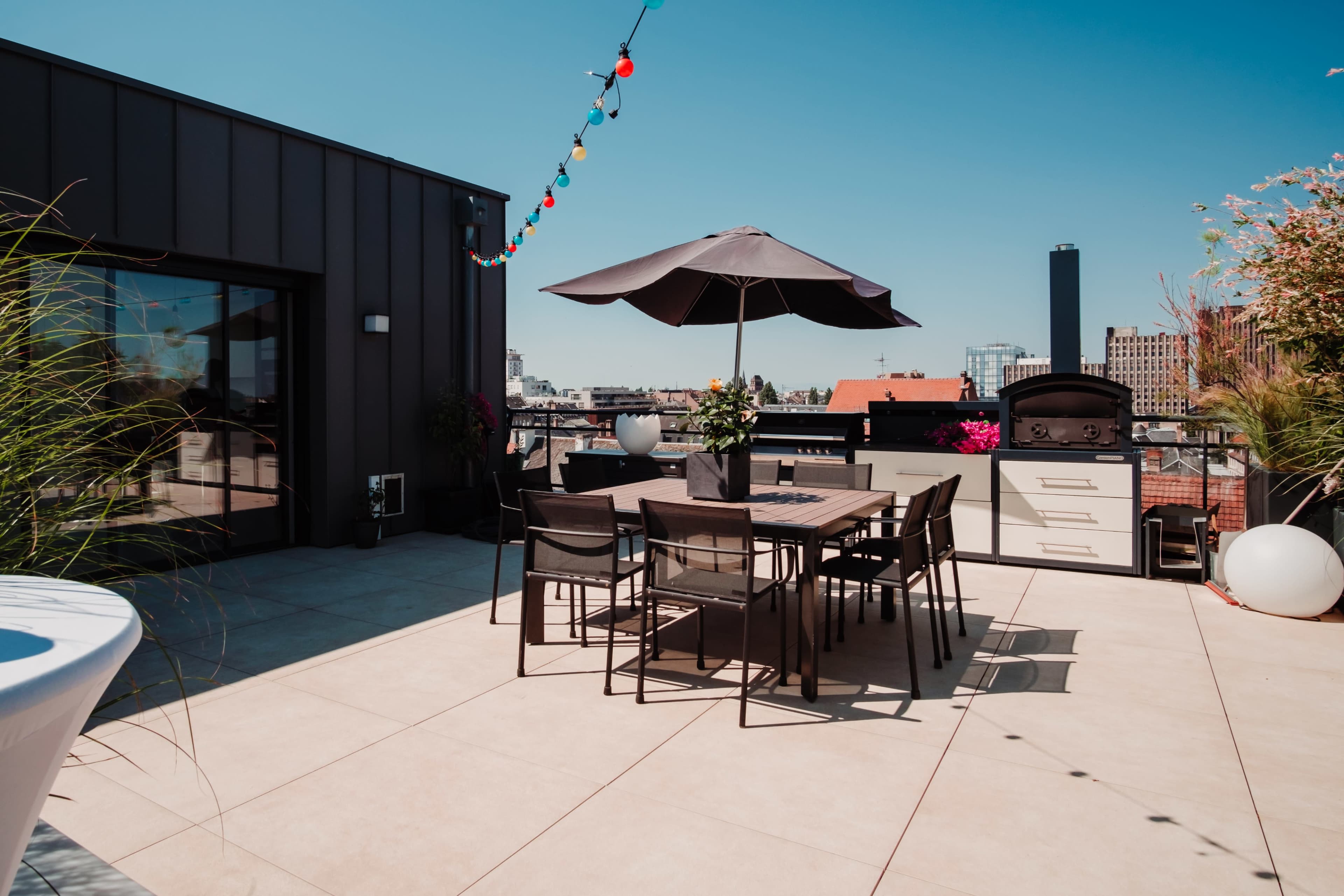 The image shows a rooftop terrace with a dining table, chairs, and a large umbrella, surrounded by planters and decorative lights against a clear blue sky.