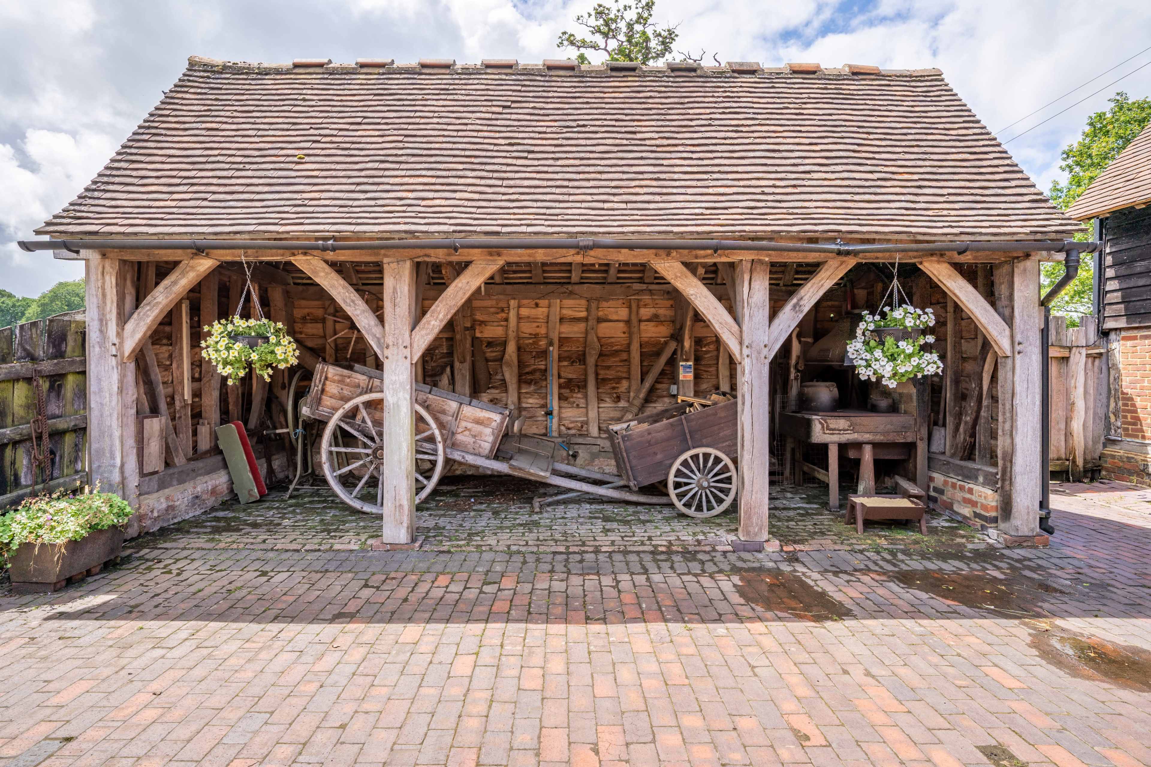 A rustic wooden barn with a covered area that houses an old wooden cart and a workbench, surrounded by potted plants.