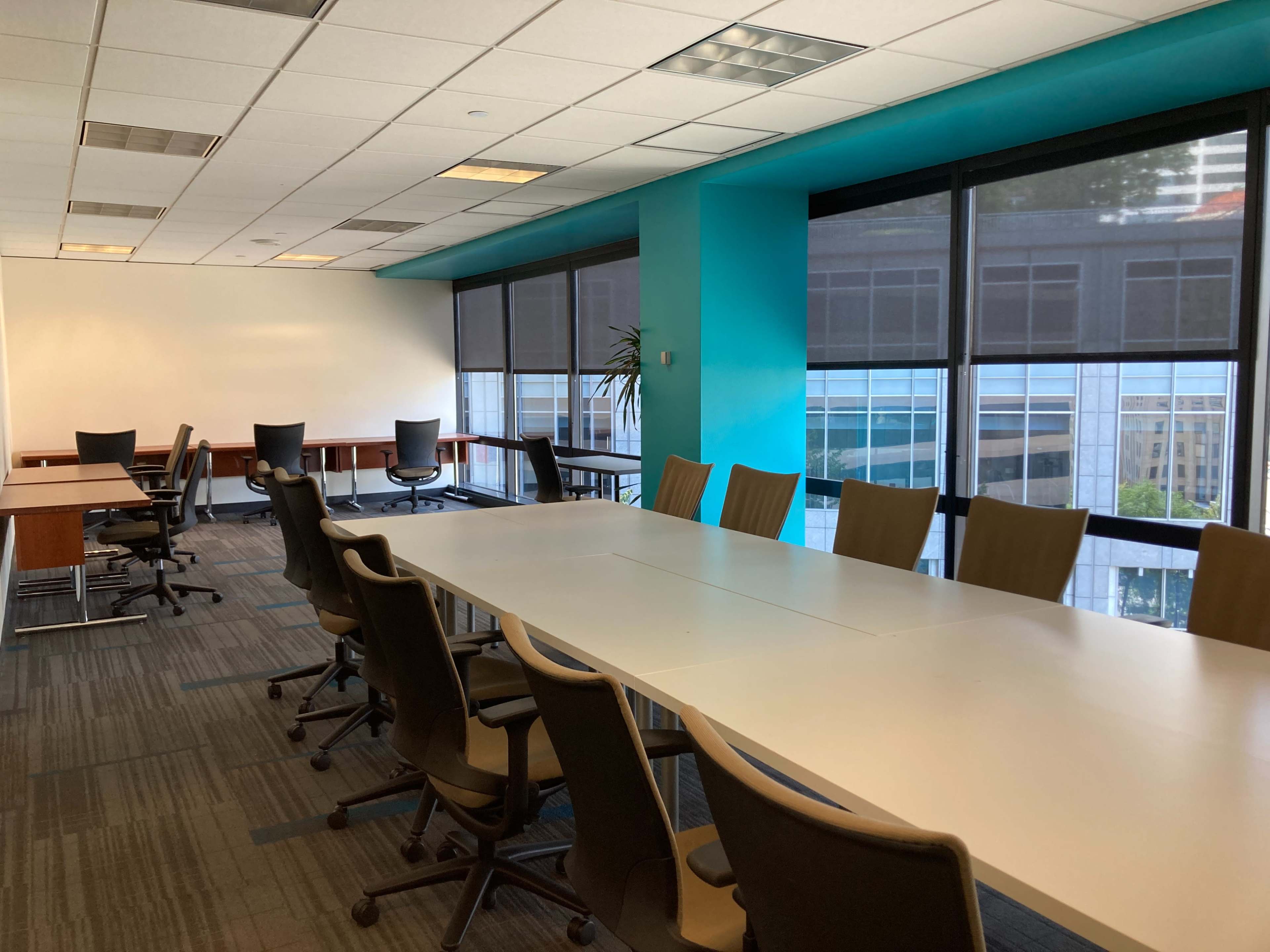 A conference room with a long white table and several office chairs arranged around it, alongside a window revealing a view of a cityscape.