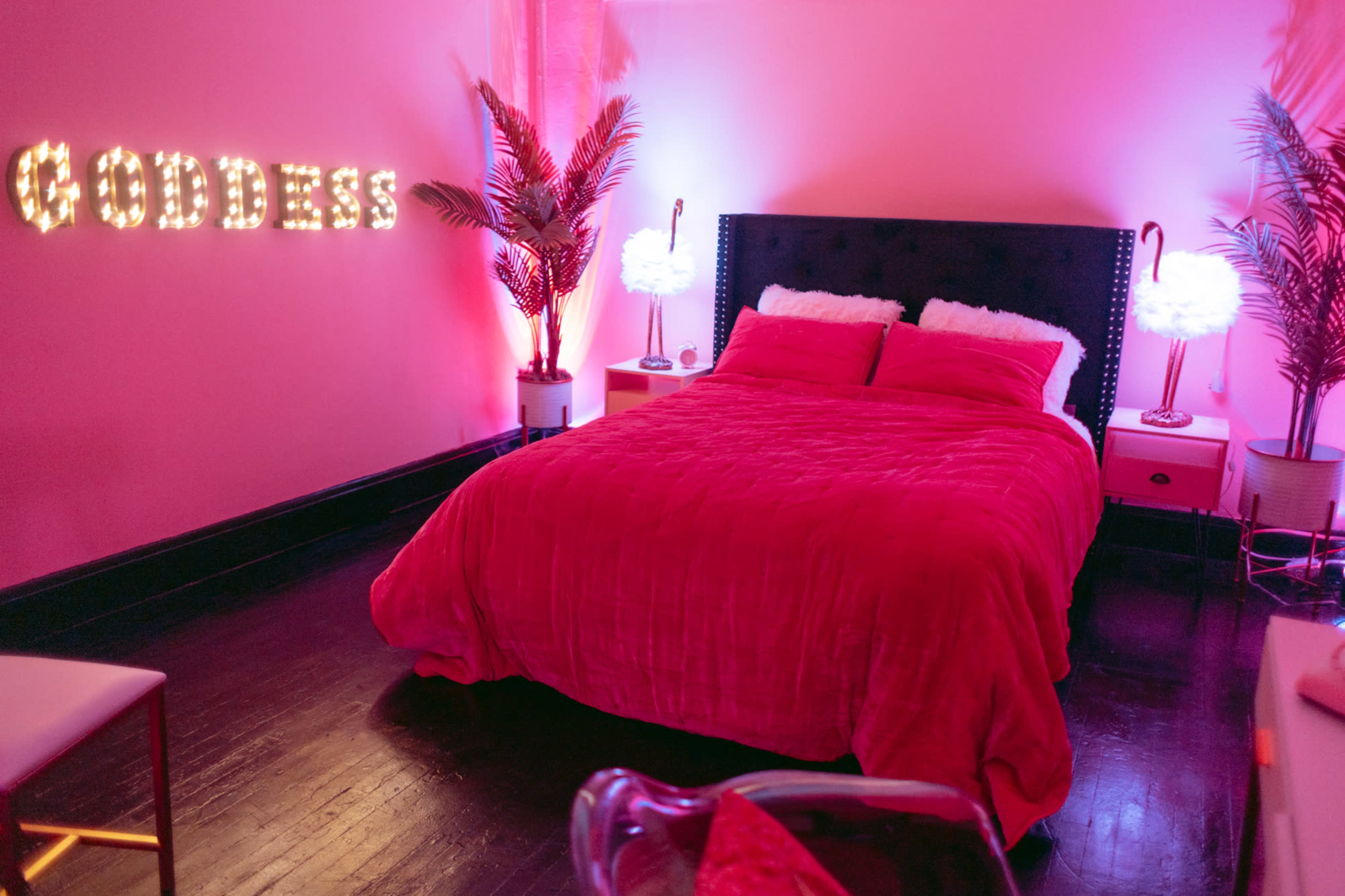 A bedroom with a pink color scheme featuring a queen-sized bed, illuminated decorative letters spelling "GODDESS," and palm plants beside light fixtures.
