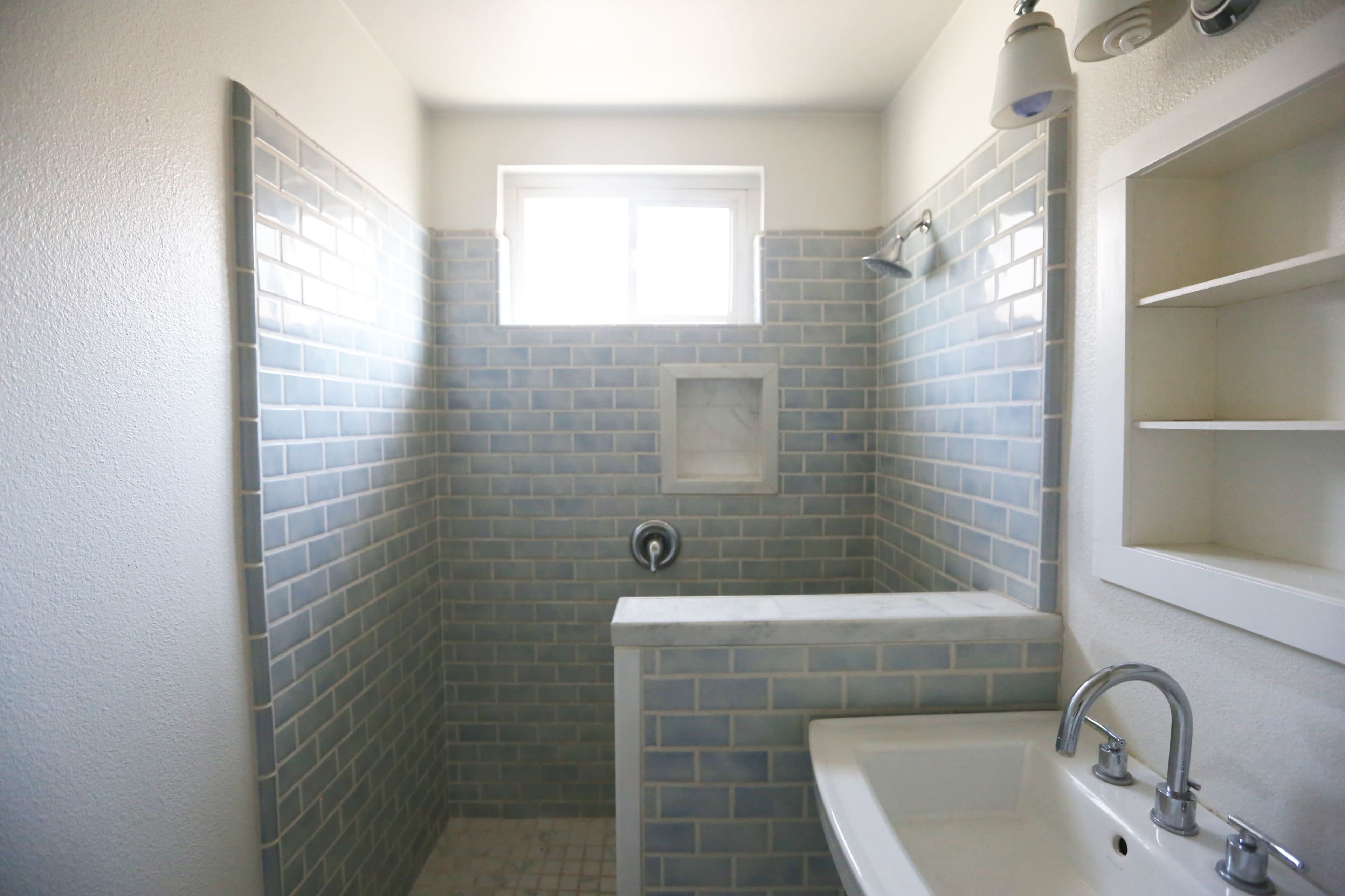 The image shows a bathroom with light blue tiled walls, a shower area with a window, and a sink counter.