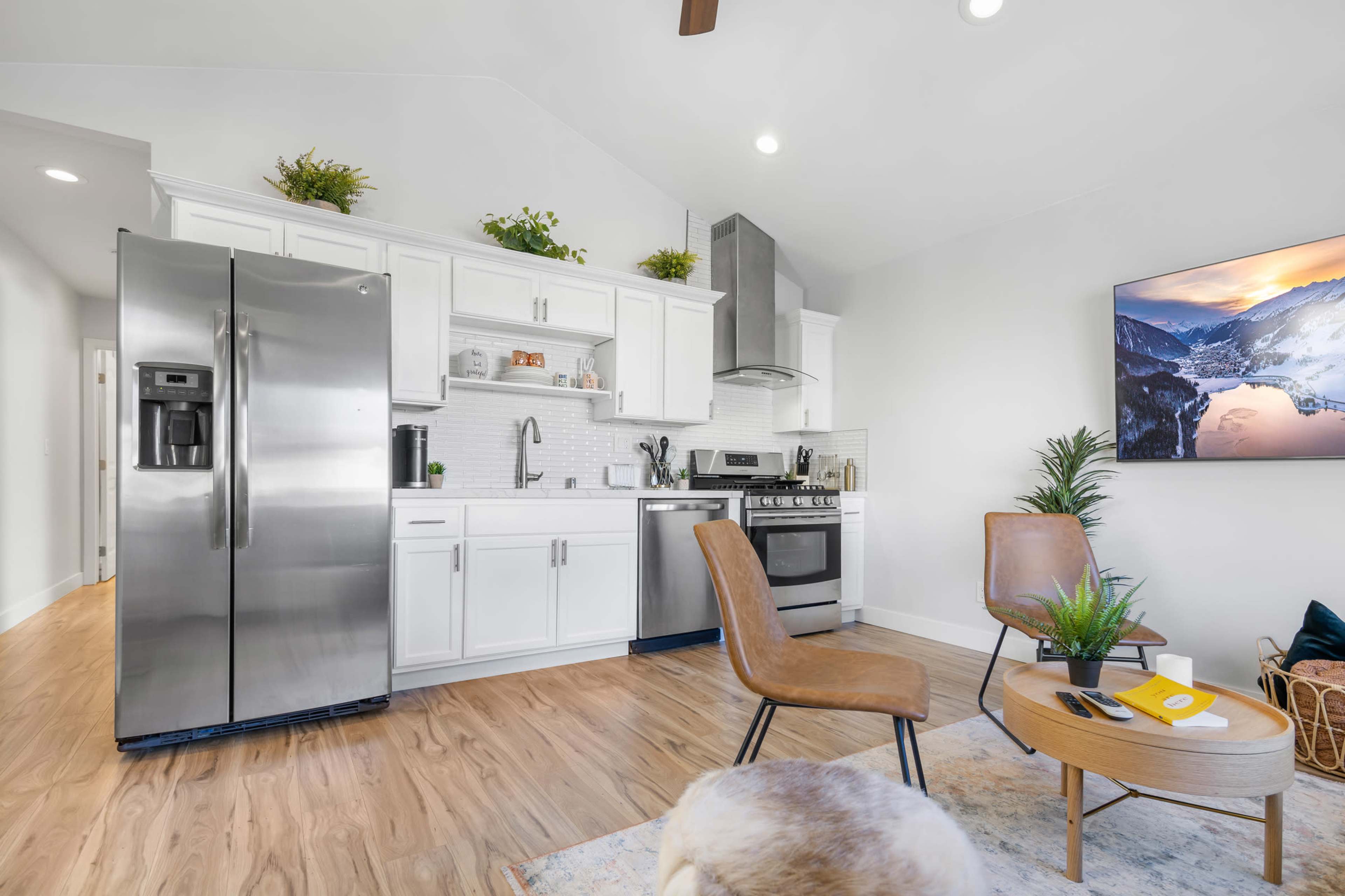 A modern kitchen and living area feature stainless steel appliances, white cabinetry, and light wood flooring, with a television mounted on the wall and two chairs nearby.