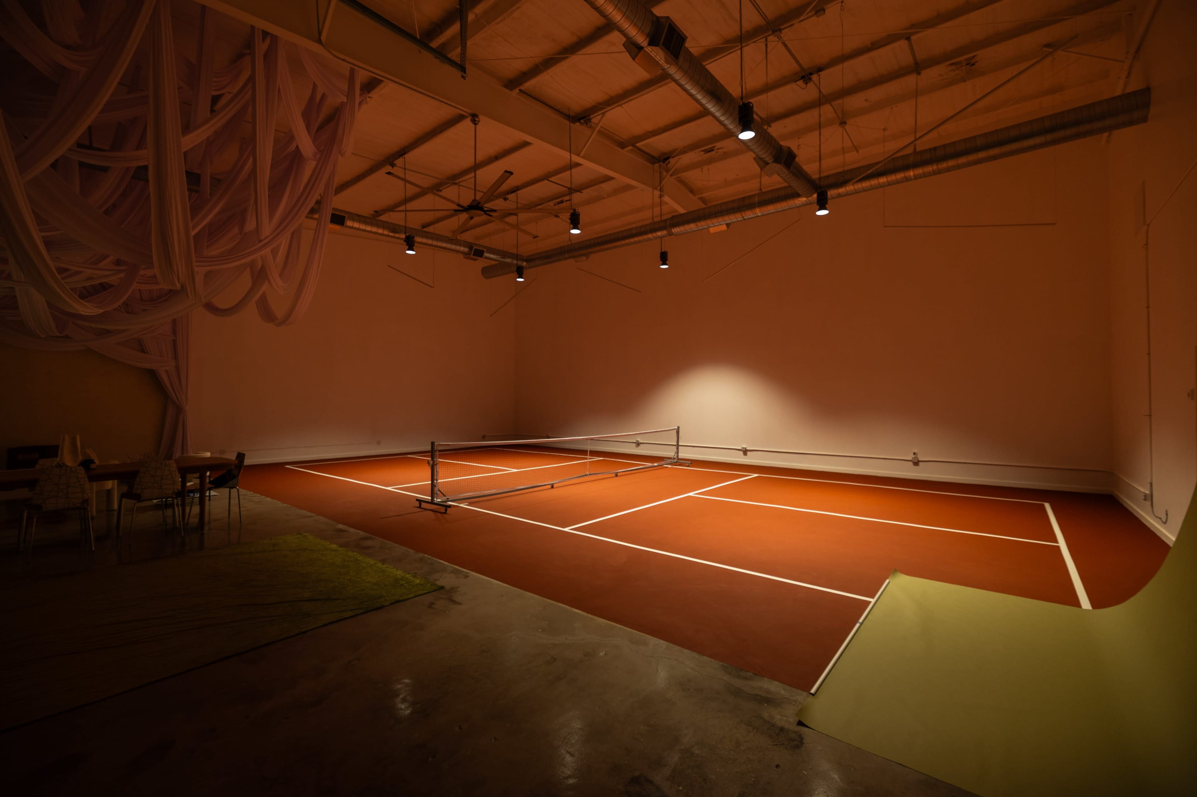 The image shows an indoor tennis court with a reddish-brown surface, surrounded by dim lighting and minimalist decor.