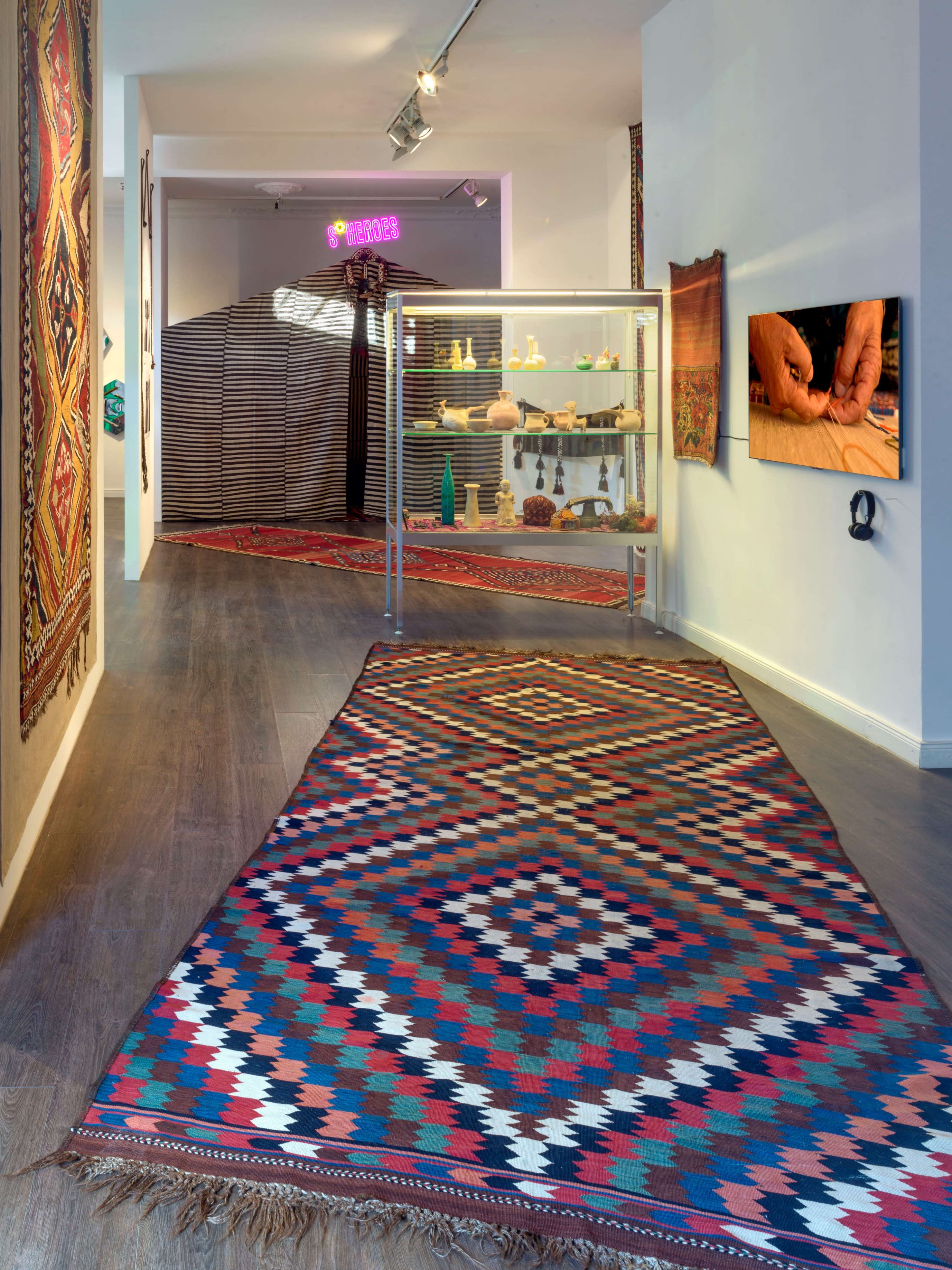 The image shows a gallery space with colorful patterned rugs on the floor, exhibit cases displaying various items, and walls adorned with woven textiles.