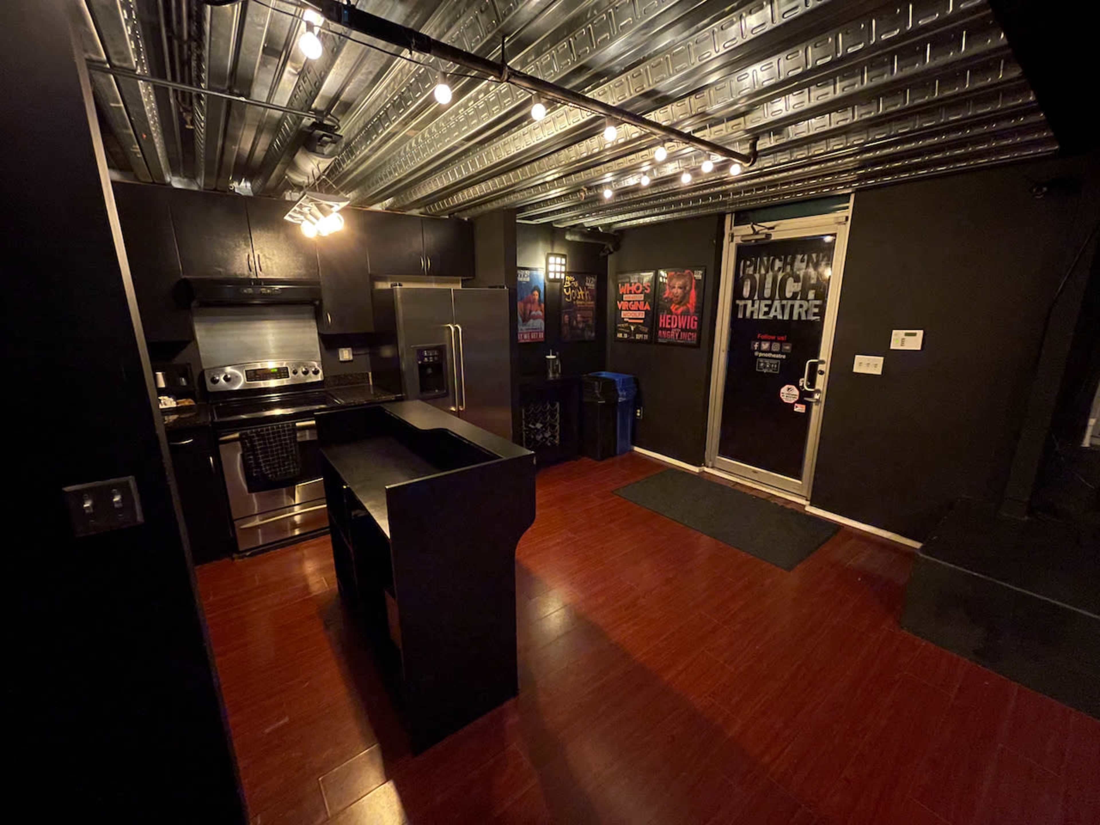 The image shows a dimly lit kitchen with dark cabinetry, stainless steel appliances, and a doorway leading to a space labeled "Buch Theatre."