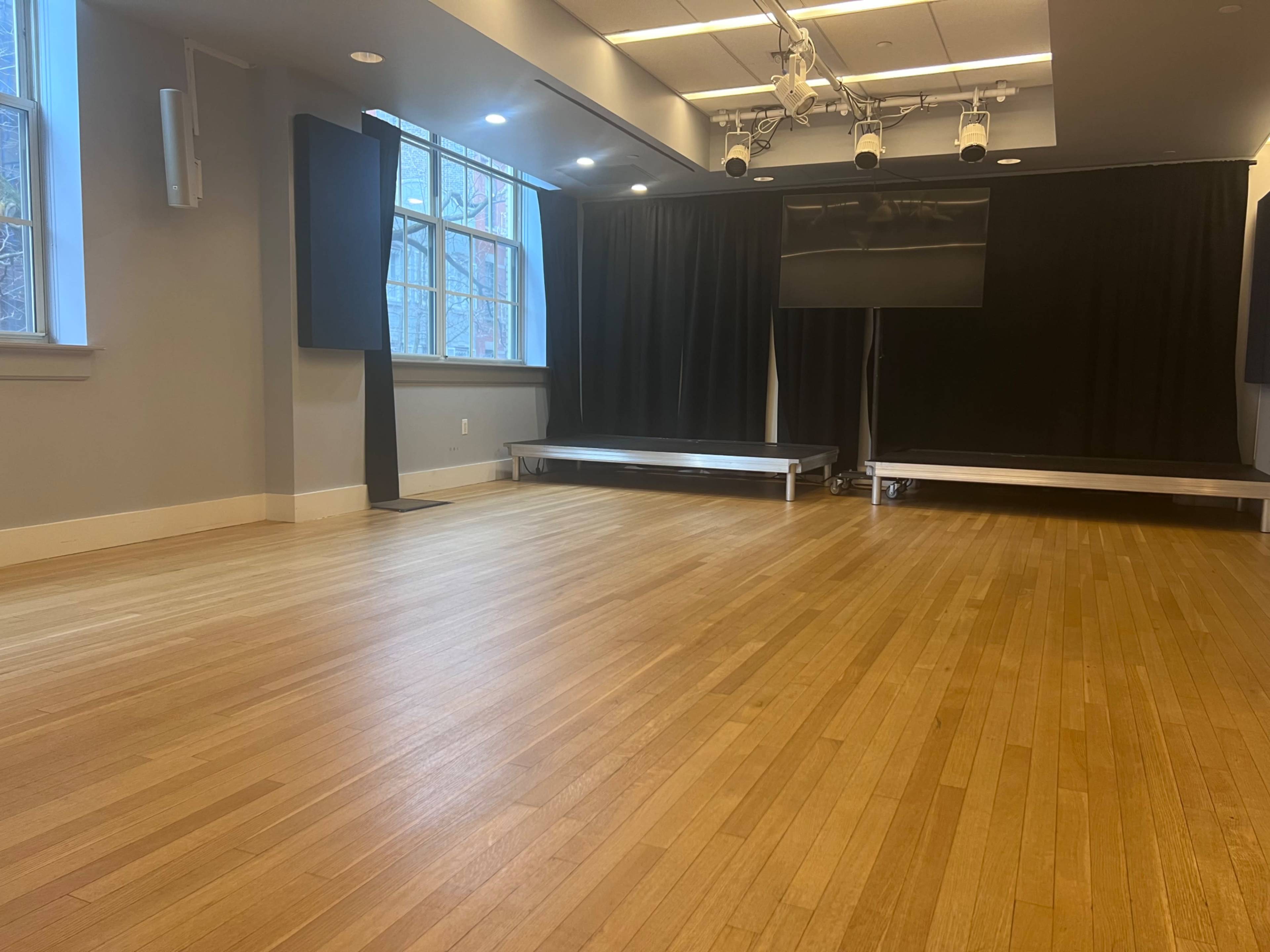 The image shows a spacious, empty room with wooden flooring, large windows, and a stage area with black curtains.