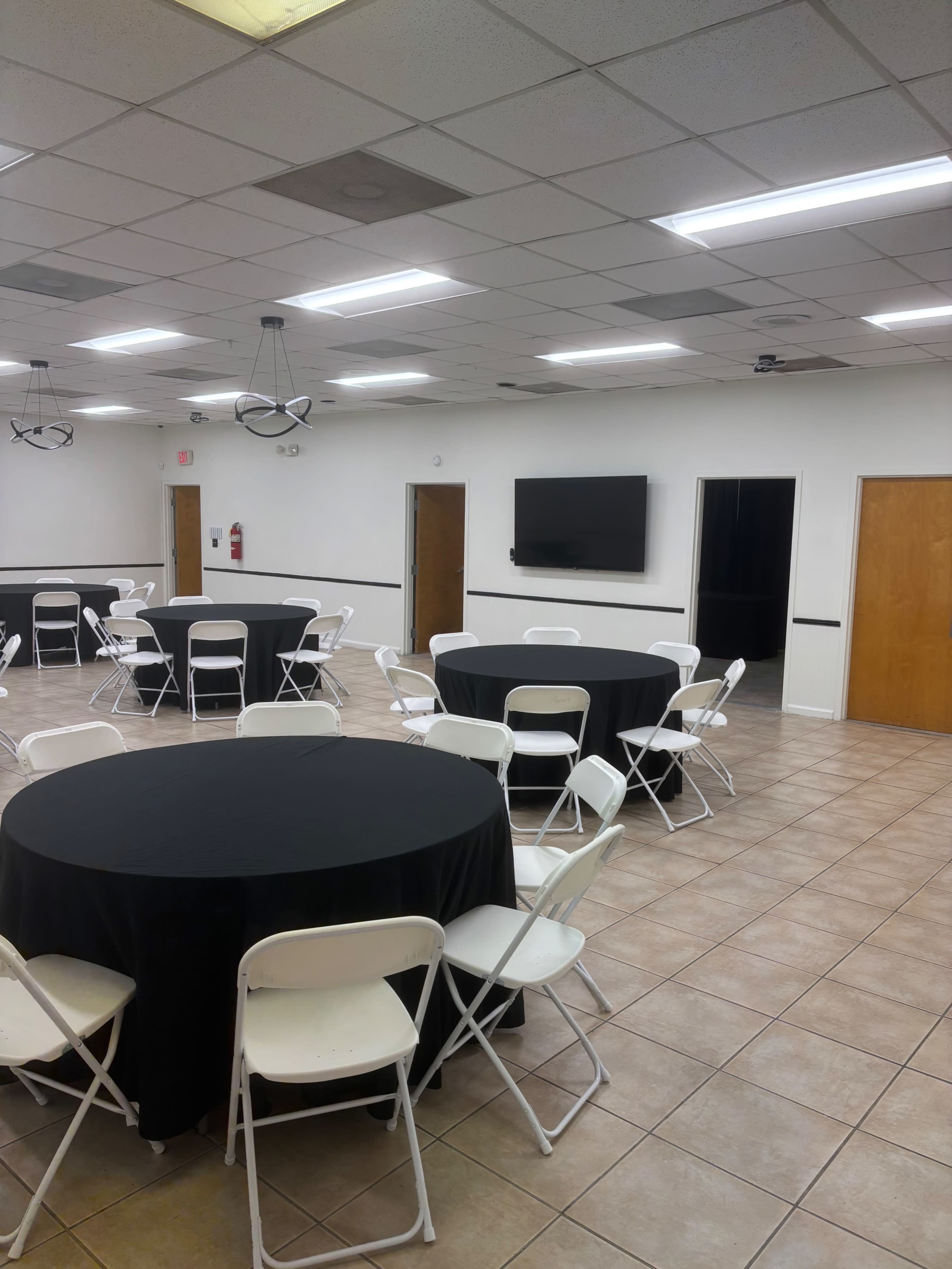 The image shows a large, empty meeting room with multiple round tables covered in black tablecloths and white folding chairs arranged around them, along with doors and a wall-mounted television.