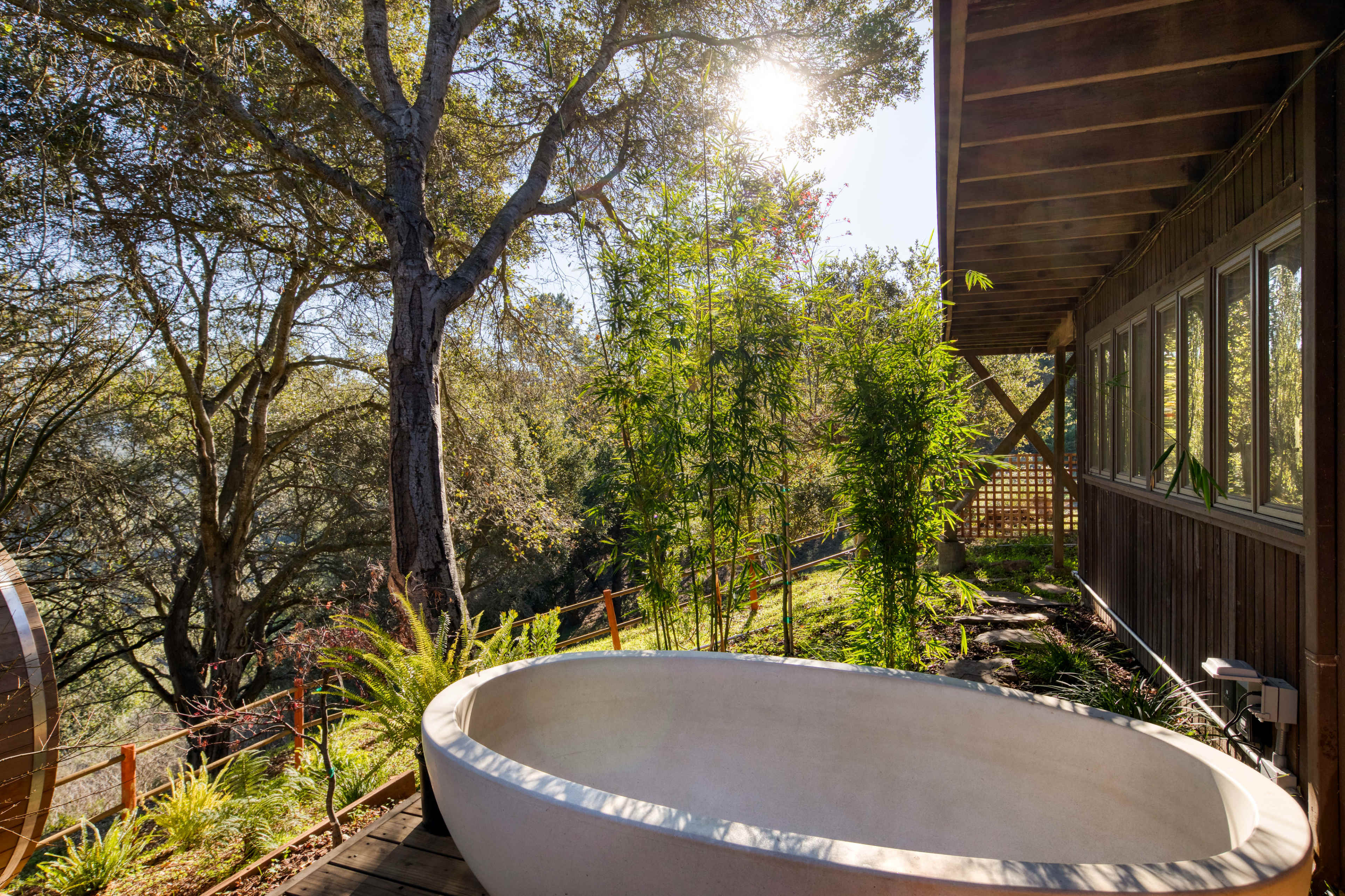A freestanding white bathtub is positioned on a wooden deck surrounded by plants and trees, with sunlight filtering through the branches.