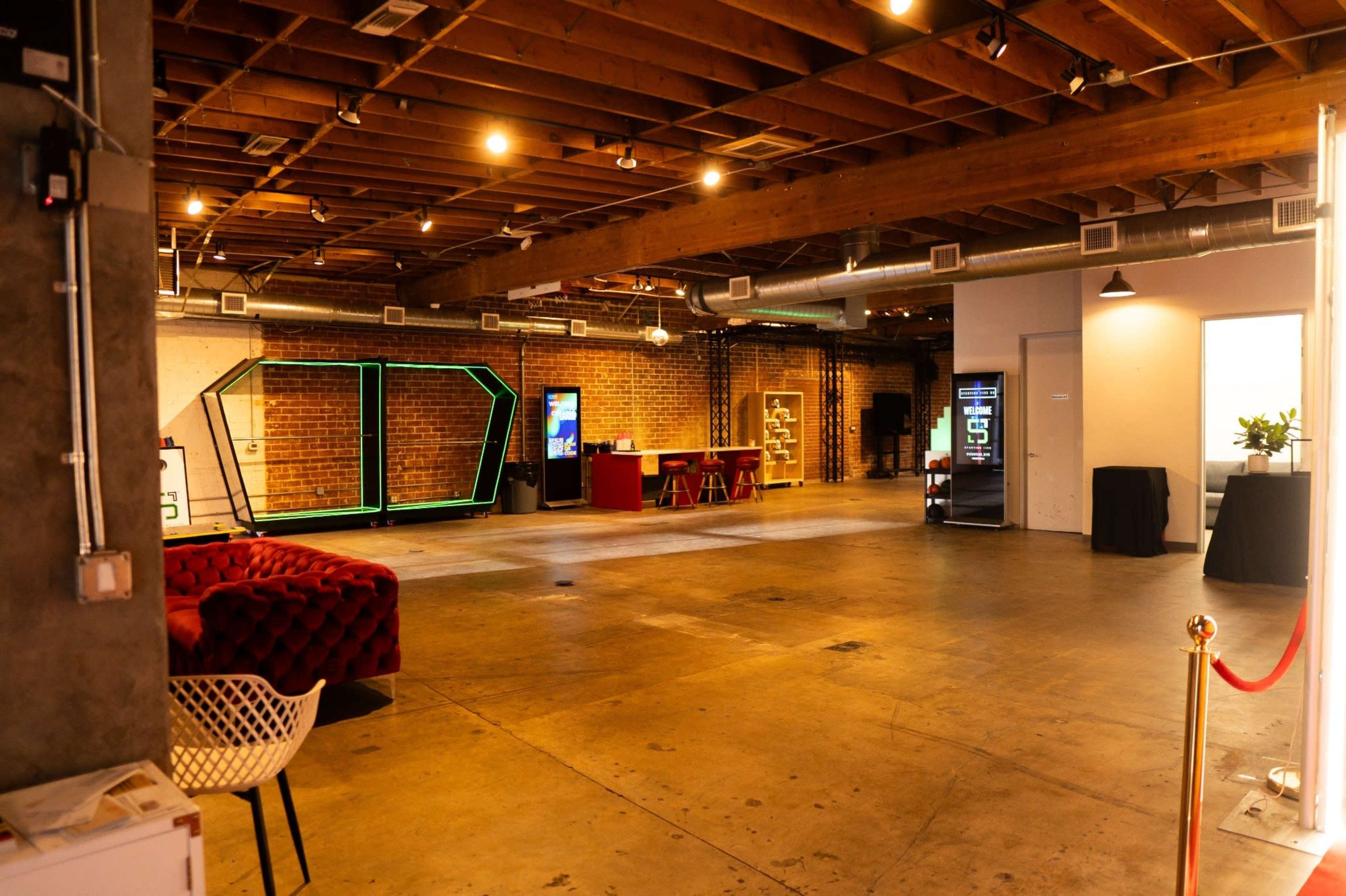 The image shows a spacious, modern event space with exposed brick walls, wooden beams, and various seating areas, including a red couch and a white chair.