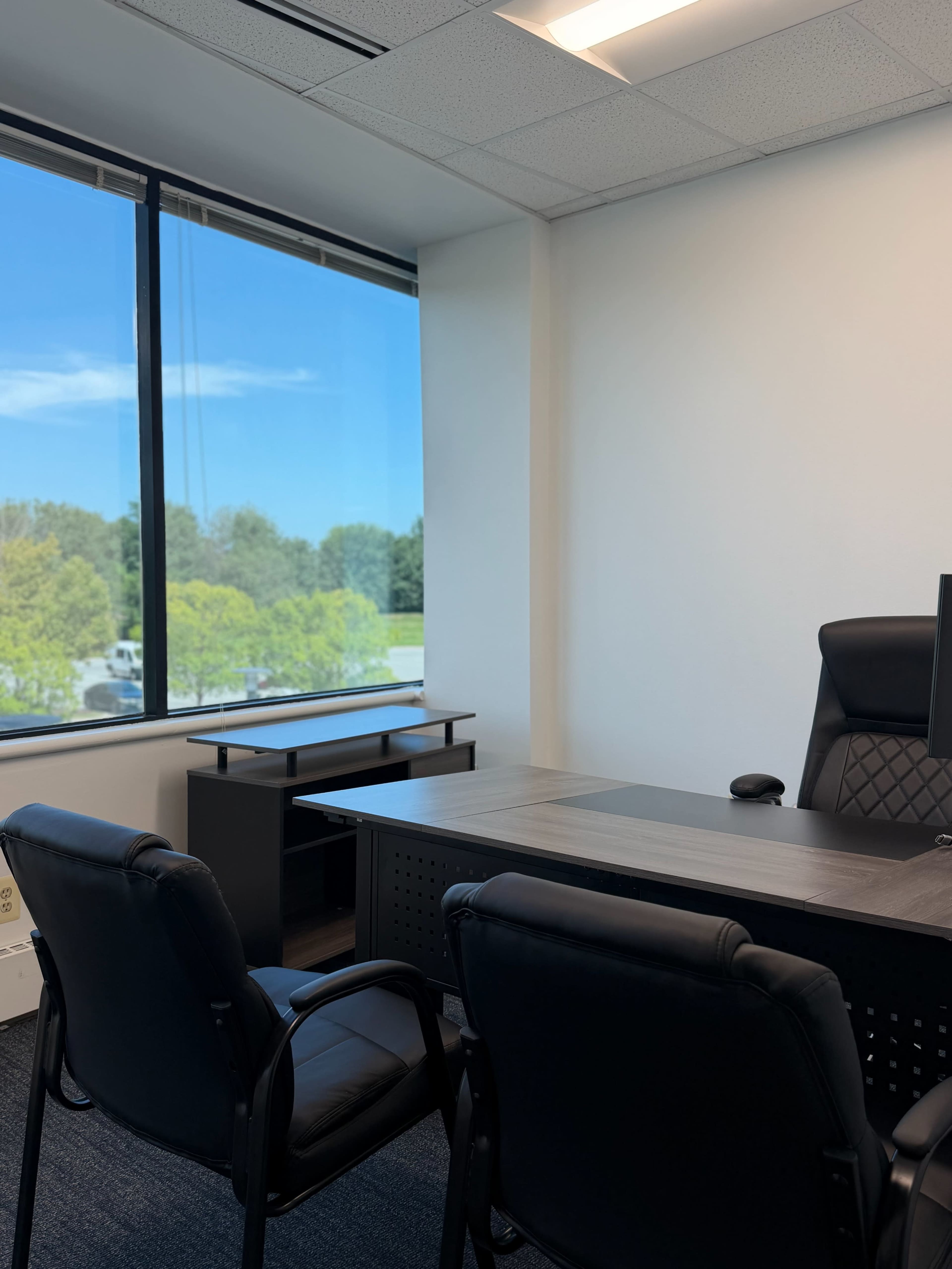 The image shows a modern office space featuring a desk, two black chairs, and a large window with a view of greenery and a clear sky.