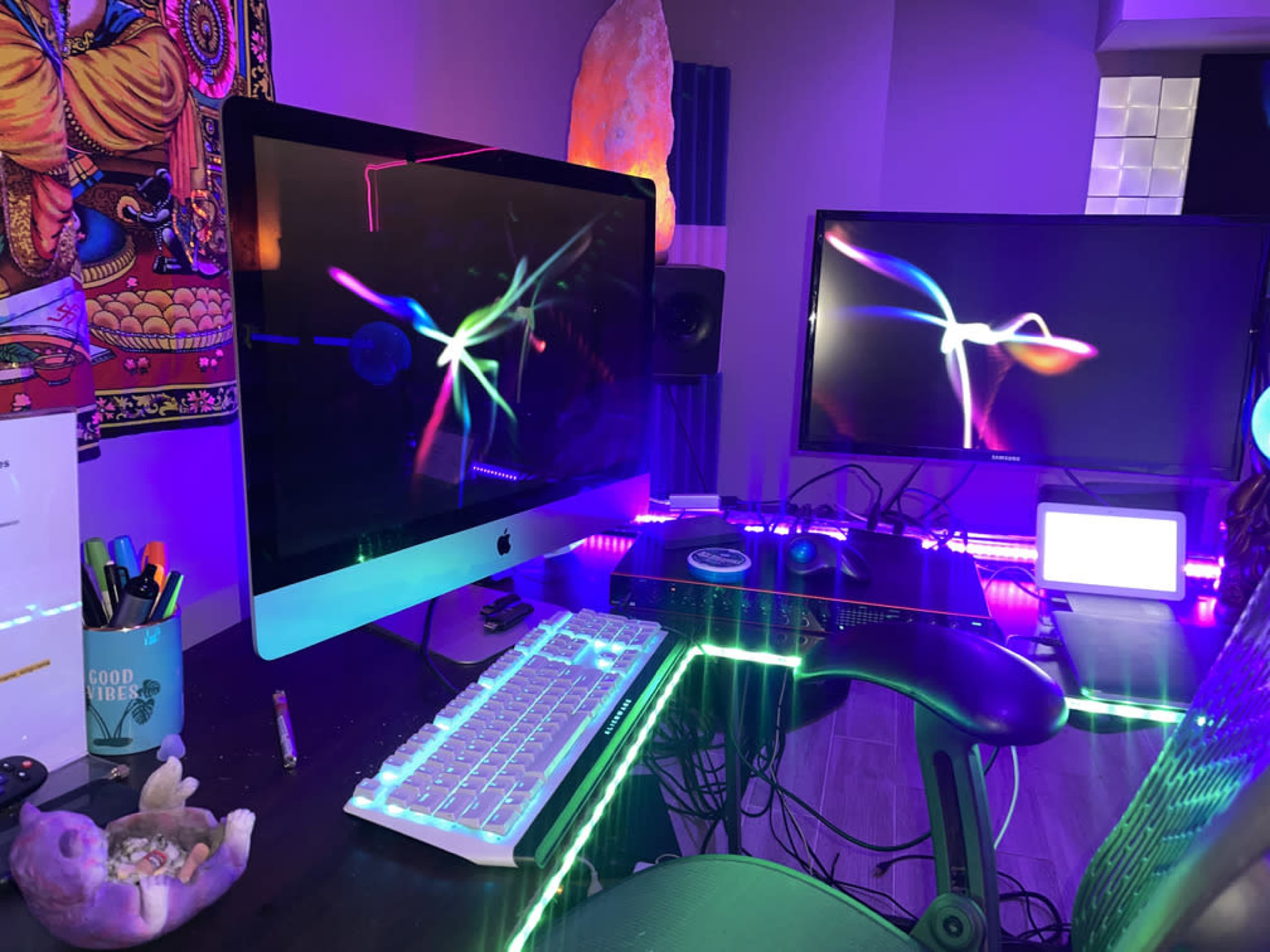 A workspace featuring two computer monitors, a keyboard, and colorful LED lighting, with a tapestry on the wall and various items organized on the desk.