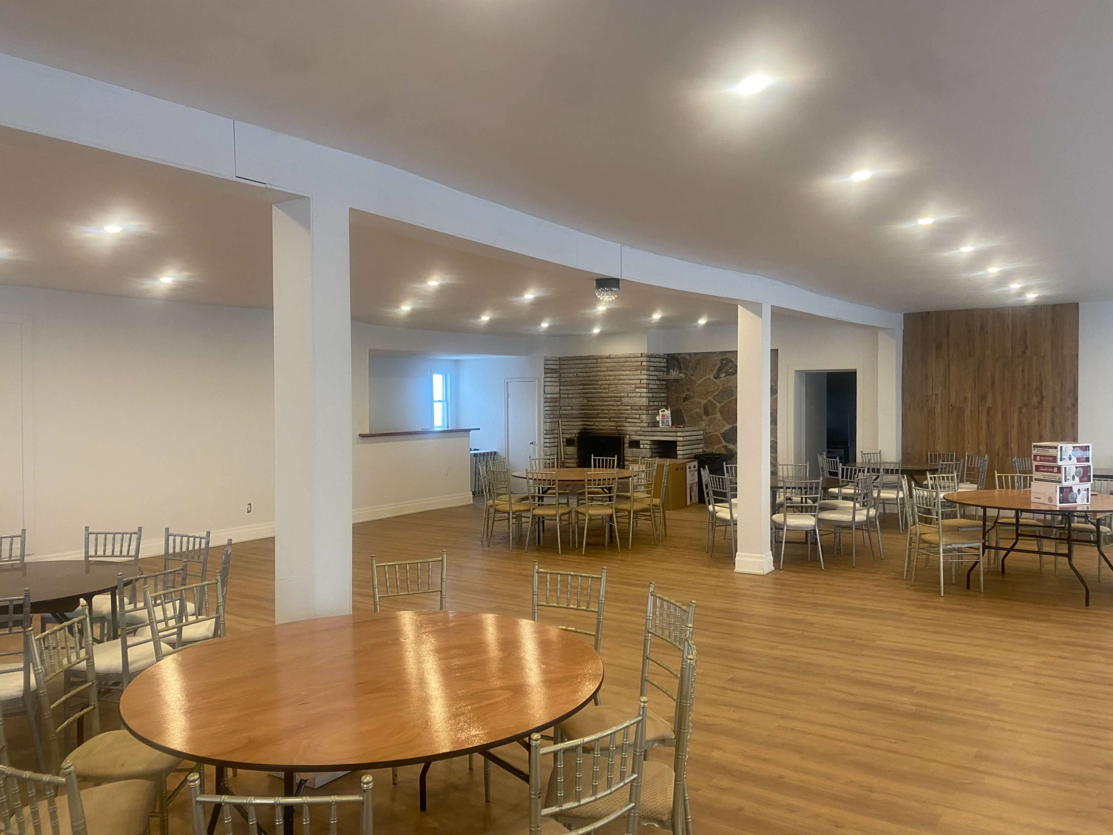 The image shows a spacious room with multiple round and rectangular tables arranged around a central area, featuring a stone fireplace and wooden accents.